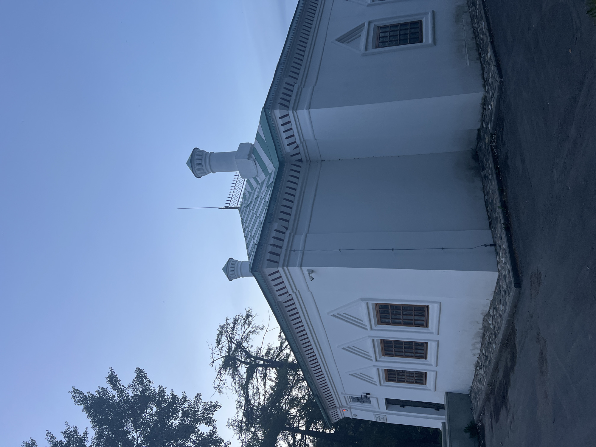 This is a full shot, exterior, eye-level, low-angle, sideways-facing photograph of a white building with a green and white striped roof, taken at dusk. The building has multiple windows with dark brown frames, some of which appear to be divided into smaller panes. There are two chimneys on the roof, each with a decorative cap. The building's exterior features decorative trim below the roofline and triangular architectural details above some of the windows. In the background, there are trees, and the sky is a clear, pale blue. The ground in front of the building is paved with cobblestones near the structure, transitioning to asphalt further away. There are no people visible in the image.