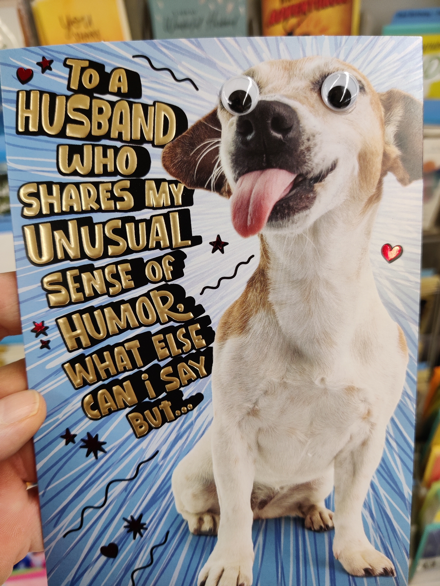 A greeting card with the text "TO A HUSBAND WHO SHARES MY UNUSUAL SENSE OF HUMOR, WHAT ELSE CAN I SAY BUT..." is being held in a person's hand. The card features a humorous image of a dog with googly eyes and its tongue hanging out, set against a blue striped background with star and wave embellishments. The context is likely a retail environment, as indicated by the blurred background of other cards and shelves. The image is captured from a close-up perspective, focusing on the card and the hand holding it.