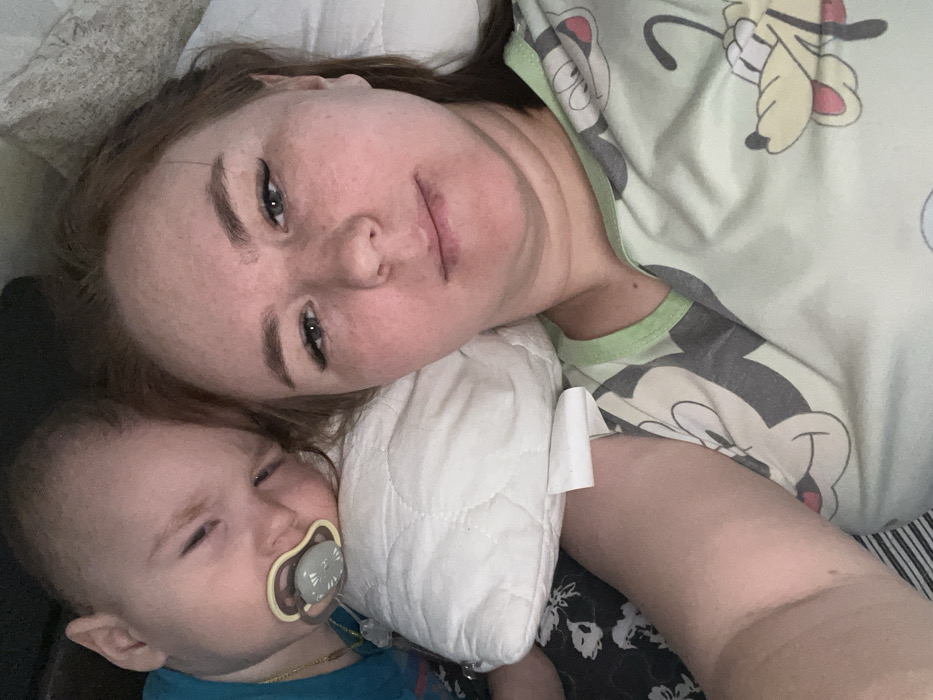 A selfie captures a woman and a baby lying on a bed. The woman, with fair skin and light brown hair, looks directly at the camera. The baby, on the left, has a pacifier in its mouth. They are both on a white pillow. The woman wears a light green top with cartoon characters. The baby is wearing a blue shirt, and a thin gold chain is visible around its neck.