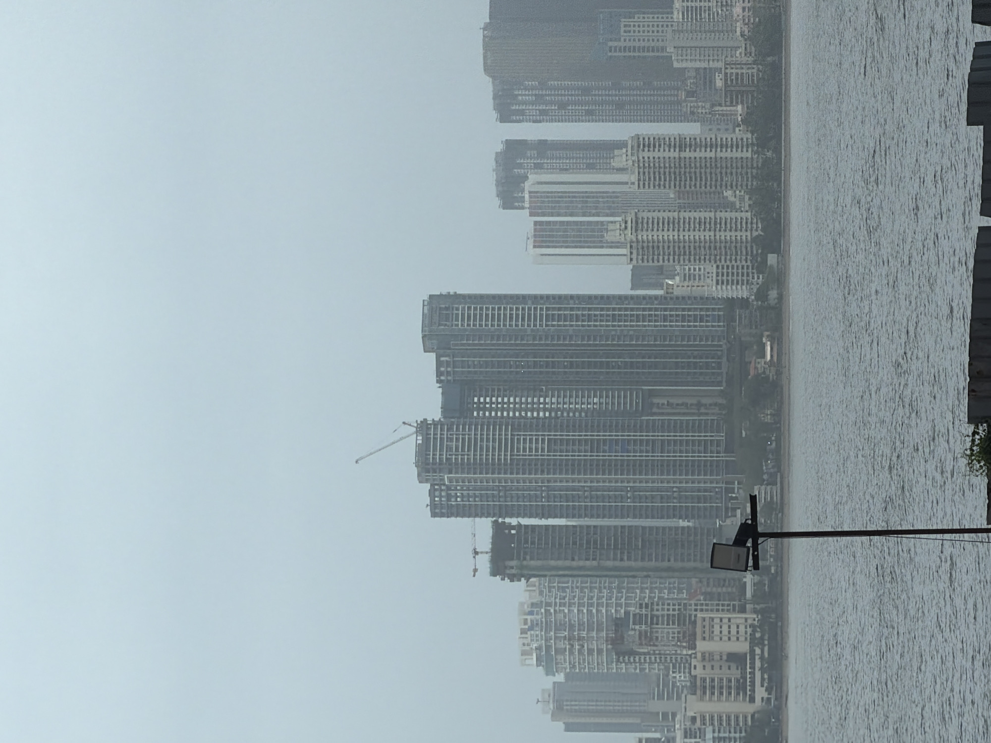 The image depicts a hazy, overcast view of the Mumbai skyline from across a body of water. The foreground features the choppy surface of the water, with a lone metal pole supporting a floodlight positioned off-center to the left. In the middle ground, the shoreline is visible, with trees and some low-lying structures interspersed between the water and the towering buildings. The background is dominated by a dense collection of modern skyscrapers of varying heights and architectural styles, some appearing to be under construction, indicated by cranes. The overall atmosphere is muted due to the cloudy conditions, which soften the details of the cityscape.