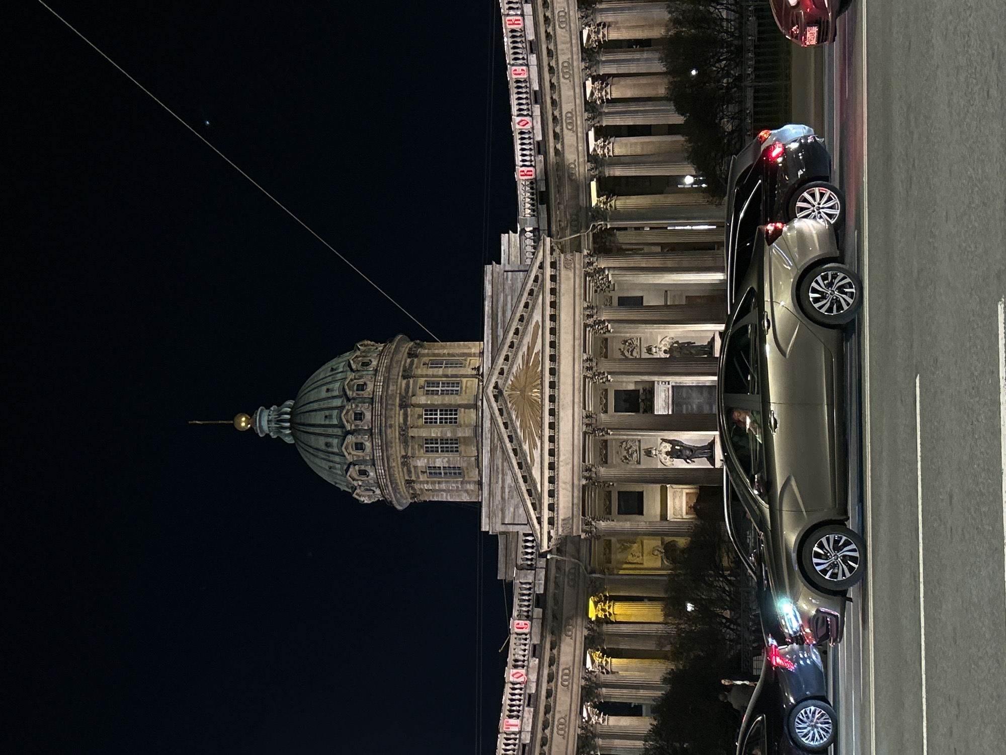 A nighttime scene in Saint Petersburg, Russia, features the grand Kazan Cathedral and a street with vehicles. The imposing classical-style cathedral, with its prominent green dome and colonnaded facade, is brightly illuminated against the dark sky. Intricate architectural details and window patterns are visible across its stone structure.
On a balcony railing across the building's front, red illuminated letters clearly spell "БЛАГОРОДНЫЙ БОГЕМА" (Noble Bohemia).
To the right of the cathedral, a paved street runs parallel, showing several cars. A light-colored sedan is positioned prominently, followed by a darker vehicle, and a red car further down the road. The cars' lights are visible, and one person appears to be in the driver's seat of the light-colored car. White road markings segment the street. A thin, light-colored wire stretches diagonally across the upper left portion of the dark night sky. The image depicts a quiet, yet active, urban evening with the historic architecture as its centerpiece.