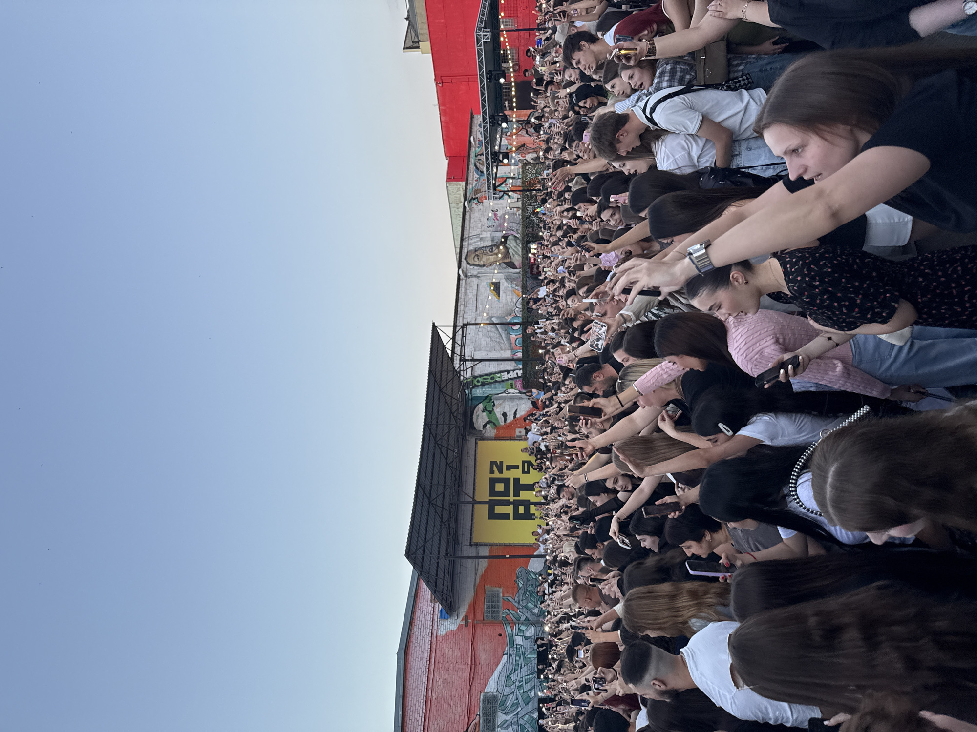 The image depicts a large crowd of people at an outdoor event, likely a concert or festival, in Krasnodar, Russia. The crowd is densely packed, with many individuals holding up their mobile phones to record or photograph the event. The setting appears to be an urban environment with industrial-style buildings in the background. One building on the left is painted with a mural, and a yellow banner with black lettering is visible in the center background. The sky is clear and pale blue. The overall impression is of a lively and engaged audience.