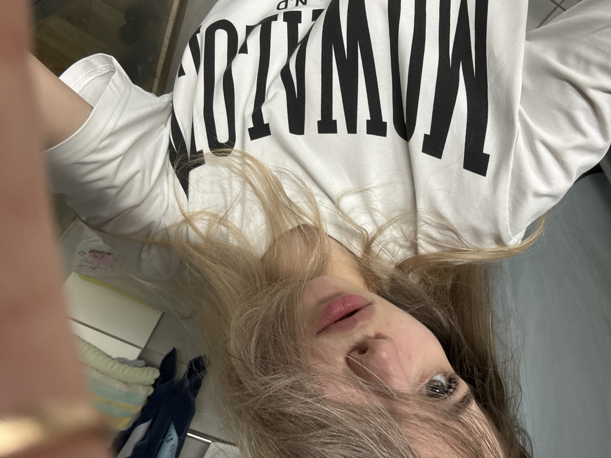 A fair-skinned woman with blonde hair is lying down, wearing a white t-shirt with large black lettering. The lettering reads "MOTIVATION". She is indoors. The exact location and the city of Chaikovsky, Russia cannot be confirmed from the image. The surrounding environment includes a bathroom area with tiled walls and various items possibly including towels and clothing.
