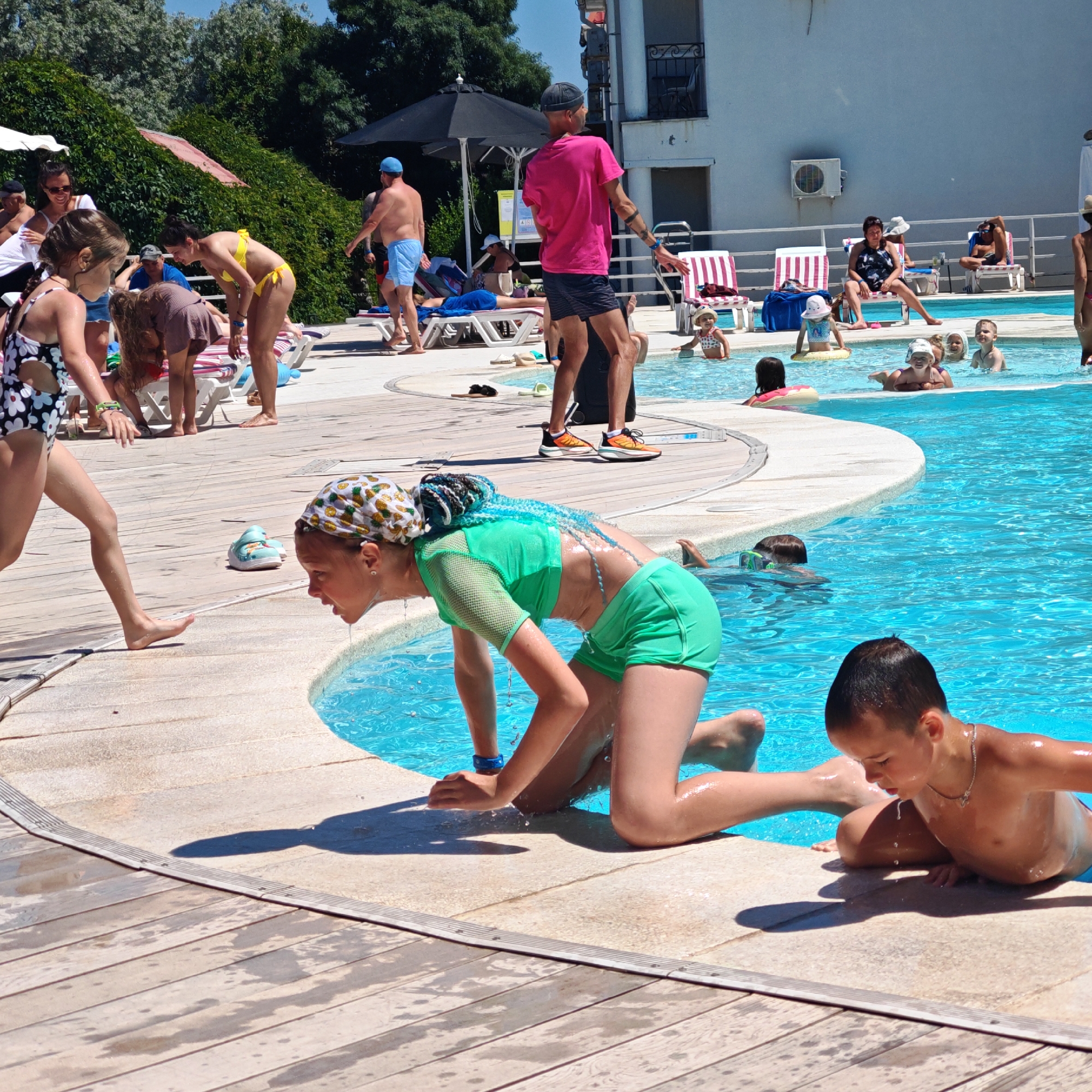 The image depicts a sunny day at an outdoor swimming pool, likely in a resort or hotel setting. The pool is filled with bright blue water, and several people are visible enjoying the water and the surrounding area. In the foreground, two children are interacting at the edge of the pool. One child, a girl with brightly colored blue braids and a green swimsuit, is leaning into the water, with water droplets visible on her face. Another child, a boy with short dark hair, is also at the pool's edge, leaning forward.
Further back, several other individuals are present. A woman in a yellow bikini is bending over near the pool. Other people are seated on lounge chairs or standing near the pool, some under umbrellas. In the background, a building with a white facade and balconies is visible, suggesting it is part of the accommodation. The ground around the pool is paved with wood and stone tiles, some of which are wet, indicating recent use. The presence of lounge chairs, umbrellas, and a building with balconies suggests this location is a leisure facility. The specific location "городское поселение Щелкино, Ukraine" is not visually verifiable from the provided image.