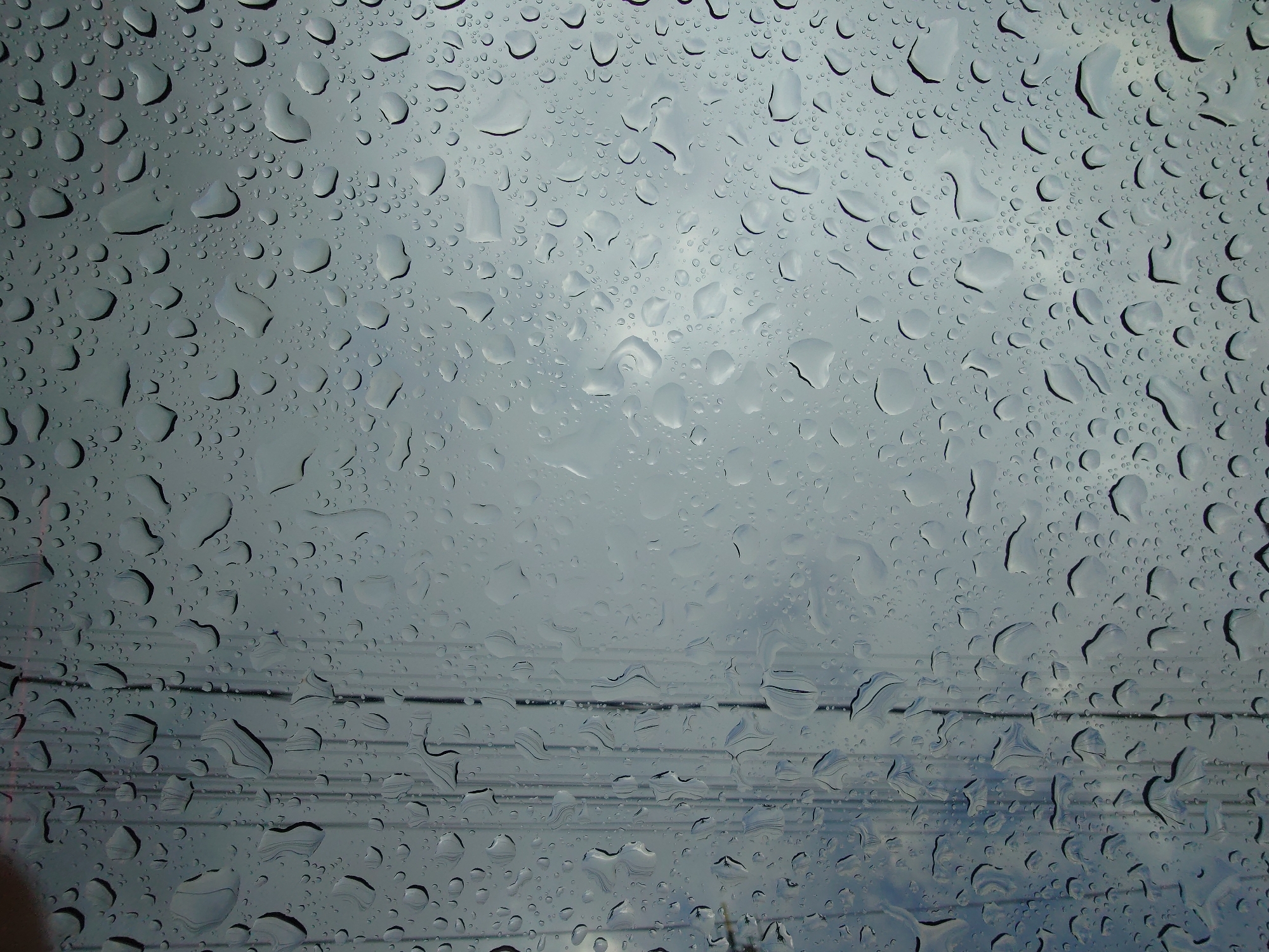 The image displays a close-up view of raindrops on a surface, likely a window or glass, with a muted, overcast sky visible through it. The raindrops vary in size and shape, with some forming larger, irregular droplets and others appearing as smaller, distinct beads. The surface is covered in a pattern of these water droplets, creating a distorted and textured visual. In the left portion of the image, faint vertical lines suggest the presence of electrical wires or some other linear structures behind the glass. The overall impression is one of a rainy day. There are no people, animals, or specific urban features identifiable in this abstract and textured composition. The location of Sokolohirsk, Ukraine, is not depicted or suggested by any visual cues in this image.
