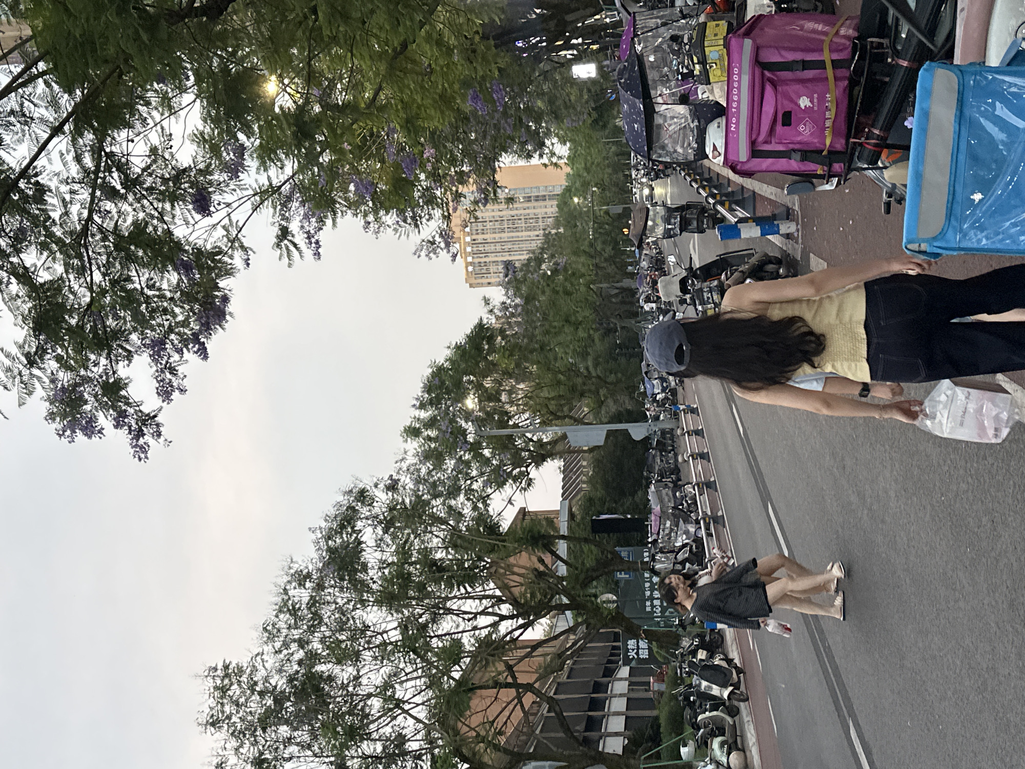 The image shows a street in the Chongqing High-tech Zone, China. A woman wearing a yellow top, dark pants, and a blue baseball cap walks along the road, holding a plastic bag. Another person is walking on the same side of the street, further down. Parked motorcycles line both sides of the street. Trees with purple flowers are visible, some branches reaching over the street. A tall building is seen in the distance.