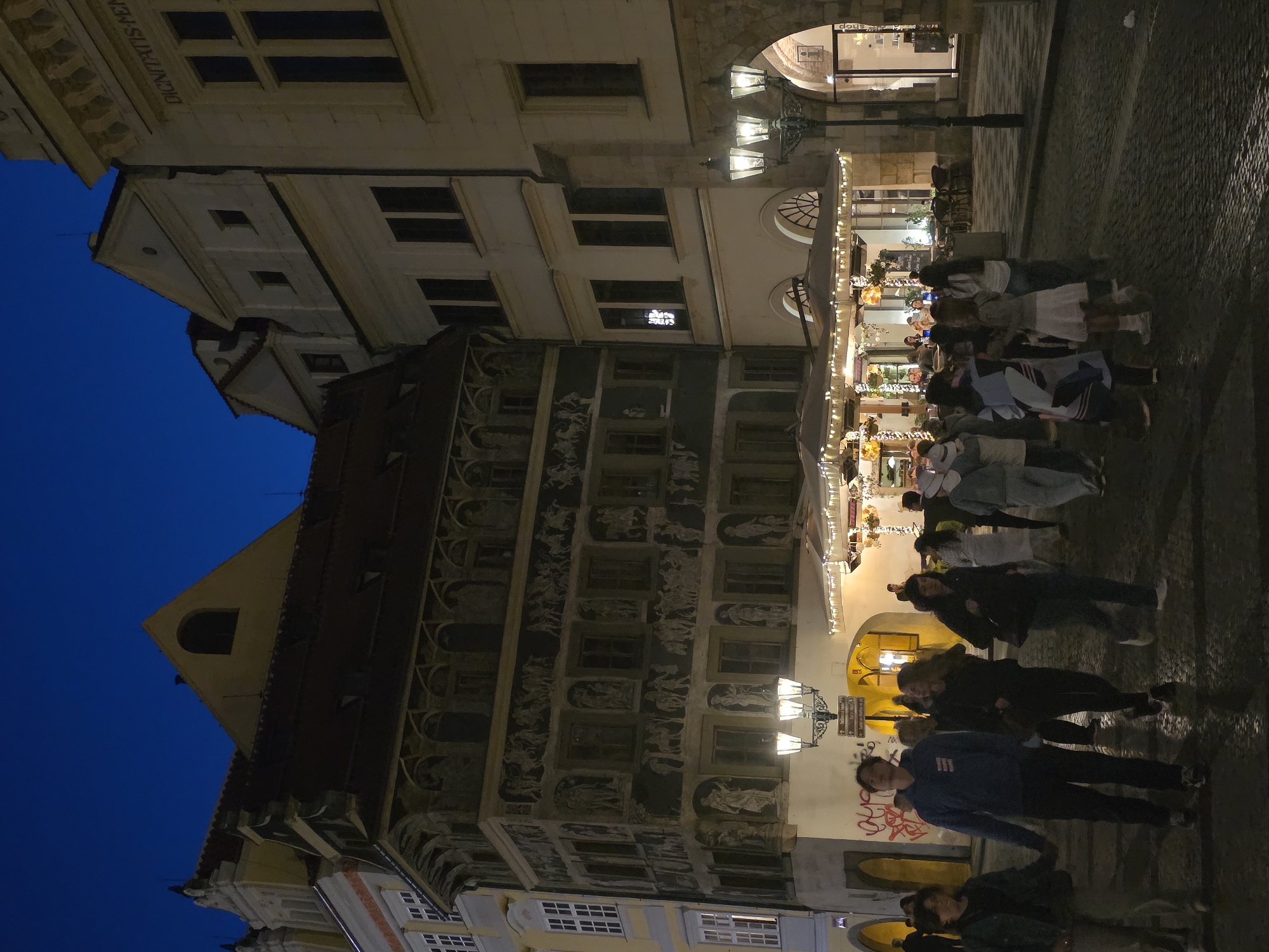 The image depicts a nighttime street scene in a European city. The architecture suggests historical buildings with ornate facades, some of which are illuminated, highlighting intricate carvings and decorative elements. In the foreground, a group of people are walking on a cobblestone street. Some individuals are dressed casually, while others appear to be tourists. They are walking in various directions, with some interacting with each other. Several lampposts light the street, casting a warm glow on the surroundings. Beyond the people, there is an outdoor dining area with umbrellas and string lights, indicating a restaurant or cafe. The sky is dark blue, suggesting it is late evening or night.