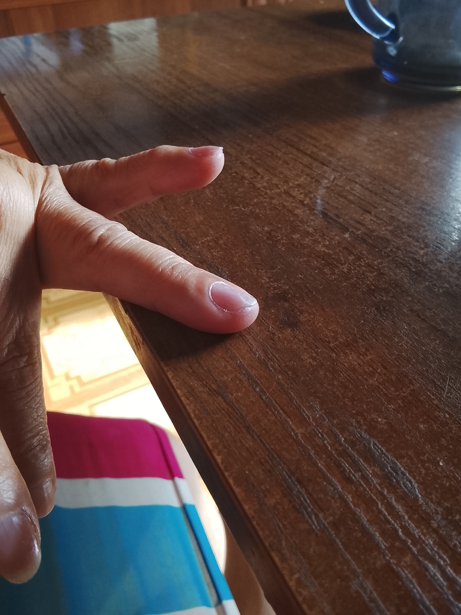 A close-up shot focuses on a person's hand and fingernail resting on a wooden table. The hand, appearing aged, is positioned with fingers gently splayed. The thumbnail, painted with a sheer, pale pink polish, shows a clean, rounded cuticle. The wooden table surface is dark brown with visible grain patterns and a slightly worn texture. In the background, out of focus, a sliver of a blue mug can be seen, and below the table, a section of fabric with horizontal stripes of pink, white, and blue is visible. The lighting suggests an indoor setting, likely during the daytime, casting soft shadows on the table. The overall mood is calm and still.