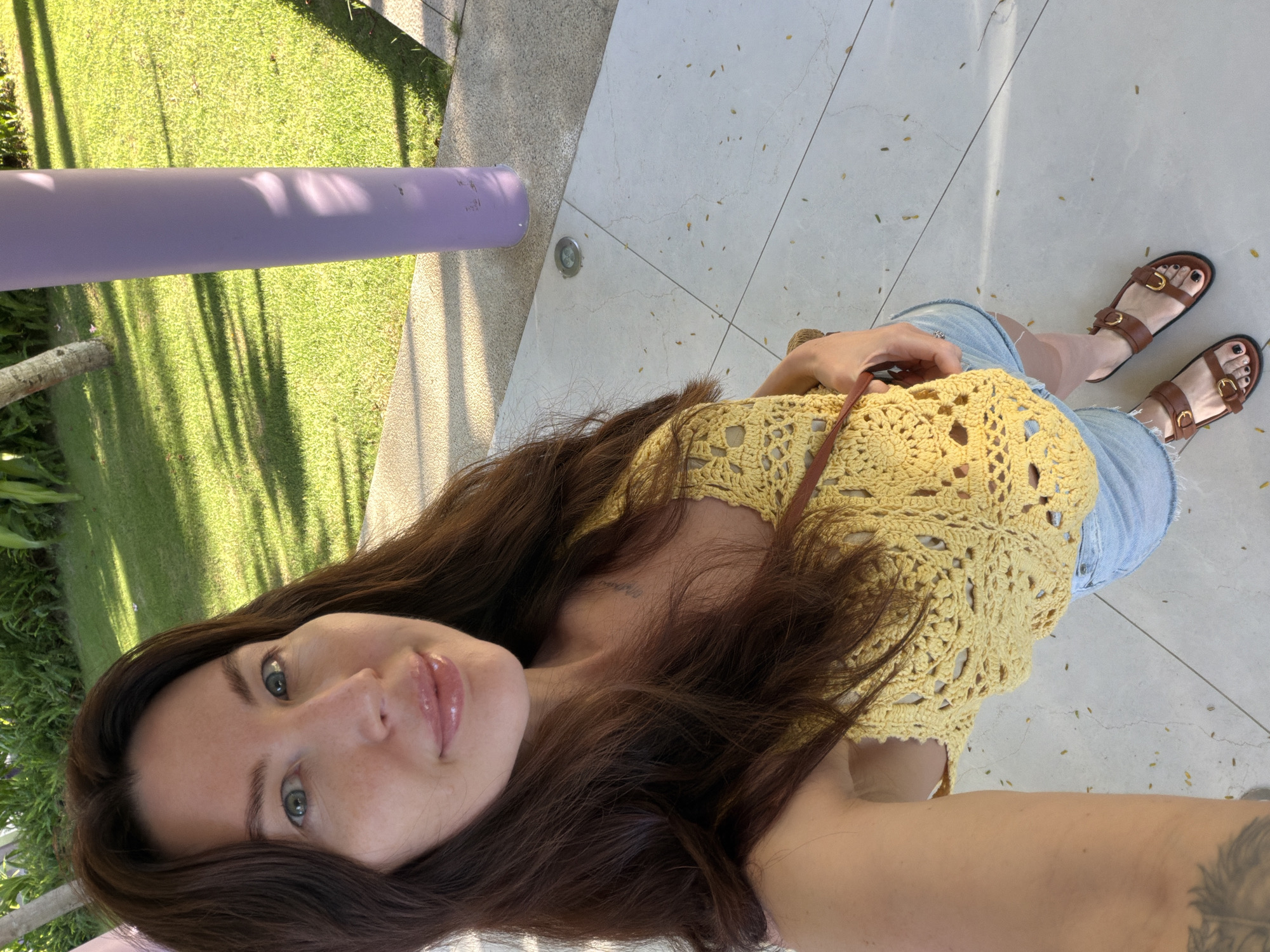 A woman is standing on a tiled outdoor surface, possibly near a building or resort, under dappled sunlight. She is wearing a yellow crochet crop top, denim shorts, and brown leather sandals. Her long, dark brown hair is down, and she is looking towards the camera with a slight smile. In the background, there is a lush green lawn, some trees, and a tall, light purple cylindrical pillar. The tiled ground has small, fallen leaves scattered across it. The scene appears to be in Dong Ninh Hoa Ward, Vietnam, suggesting a tropical or subtropical climate.
