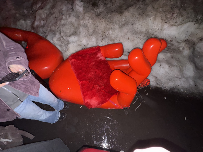 The scene depicts a large, bright red, glossy sculpture resembling a stylized human hand, lying on its side on dark, wet pavement. The palm area of the hand features a patch of red fur or plush material. To the left of the hand, another large, smooth, red, teardrop-shaped sculptural component is visible. The foreground and midground consist of dark, wet ground with standing water and small debris. In the background and along the right side, a substantial pile of dirty, melting snow is present.
A person is partially visible on the left, wearing light blue jeans, dark shoes, and a purple or maroon jacket. Their right hand holds a black smartphone. Another person's leg, clad in dark pants, is partially visible at the bottom center of the frame. The lighting is dim, suggesting nighttime or low-light conditions, with the red sculptures appearing illuminated. The location is the city of Moscow, Russia, likely near the GUM department store on Red Square, where similar "Hands" art installations by Florentijn Hofman were exhibited.