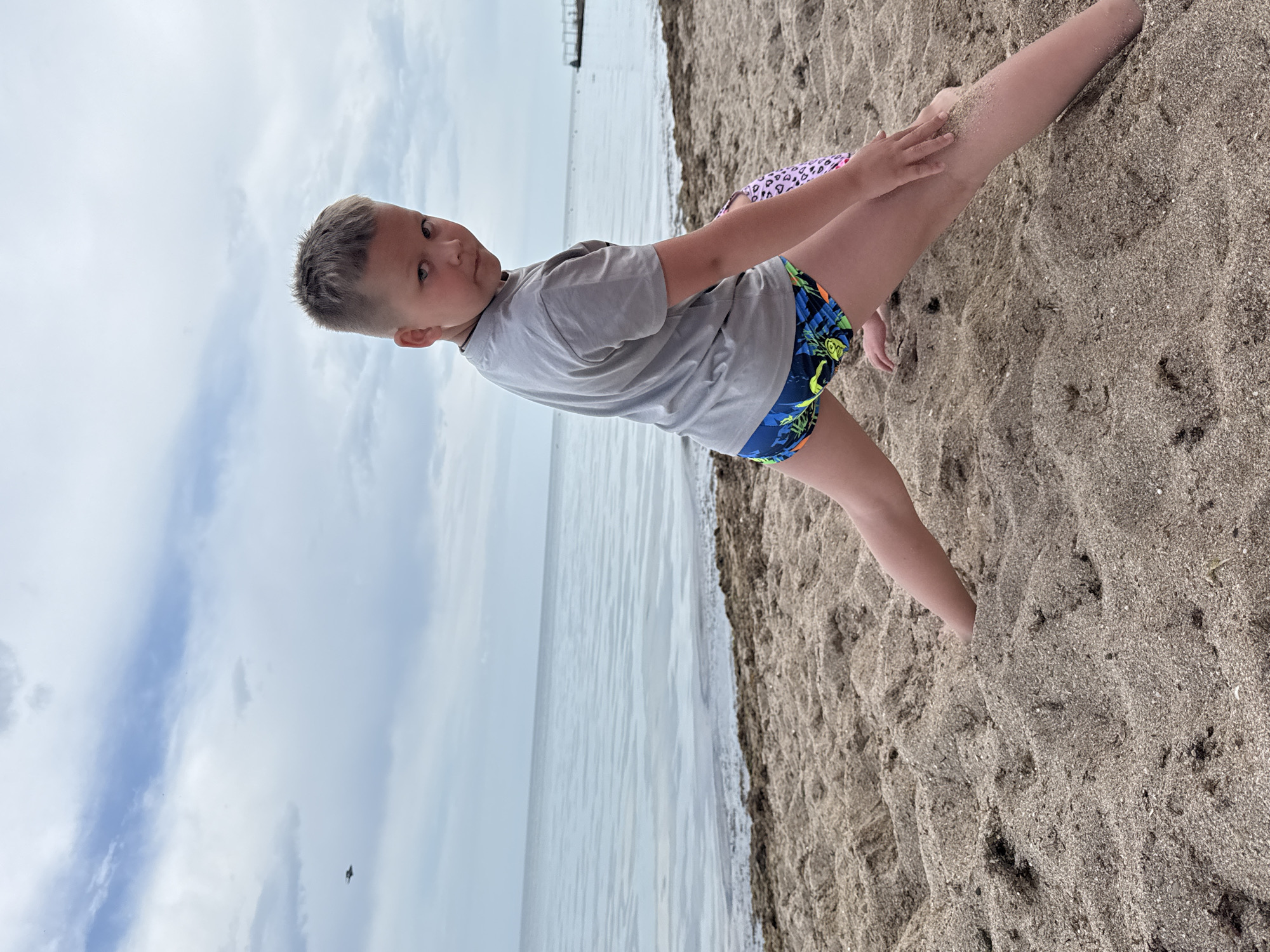 The image depicts a child at a beach in Yevpatoriya, Ukraine. The child is partially buried in the sand and wearing a gray shirt and patterned swim trunks. The sea and cloudy sky are visible in the background. There are docks on the water, and a bird is flying in the sky.