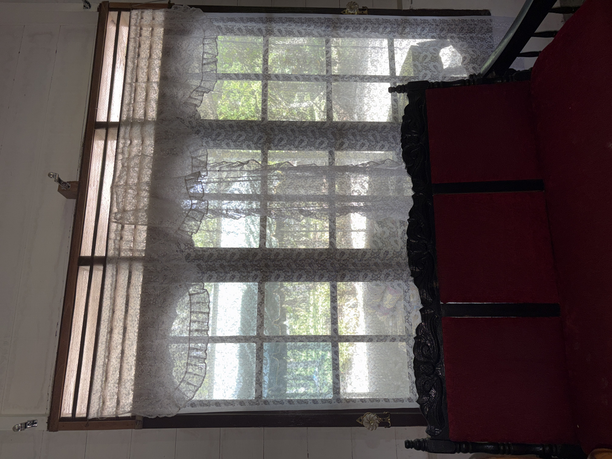 The image shows an interior scene with a window covered by lace curtains. The window is divided into multiple panes by wooden mullions, and through the curtains, the greenery of trees and sunlight can be seen. Below the window is a dark wooden bench with a red cushioned seat and backrest. Decorative carvings are visible on the headboard of the bench. A security camera is mounted on the wall above the window on the left side of the frame. The room appears to be dimly lit, with sunlight streaming through the window. The style of the window and the decorative elements suggest a possibly older building, and the scene could be a living room or a common area in a residential property in Beunyot, Indonesia. There are no people visible in the image.