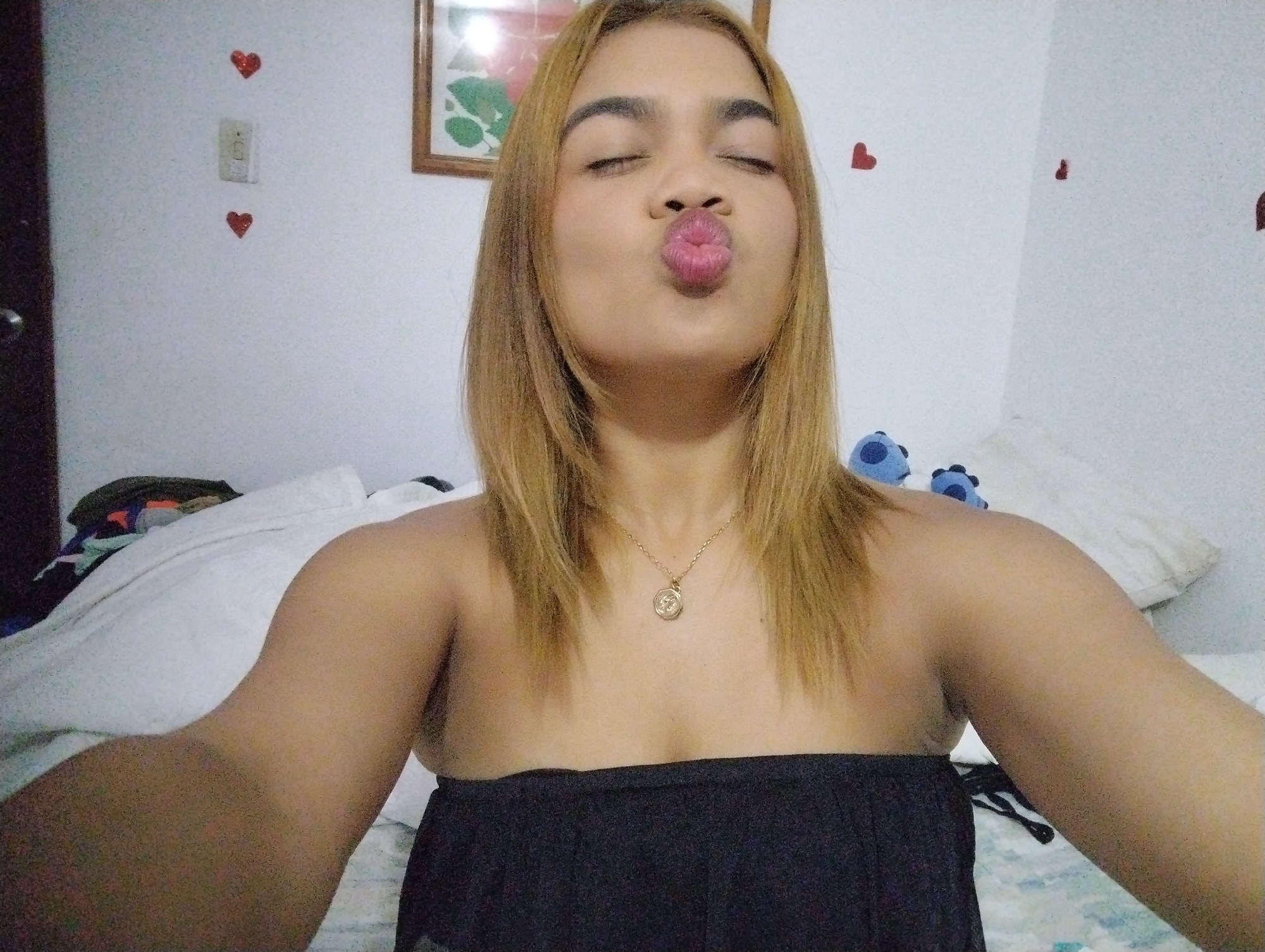 A young woman with blonde hair and a gold necklace is taking a selfie in a bedroom. She is wearing a black strapless top. She is making a kissing face with her eyes closed. The bedroom has white walls decorated with small red heart stickers. There is a framed picture on the wall behind her. The bed has white sheets and there are some items on it. The location is Porlamar, Venezuela.