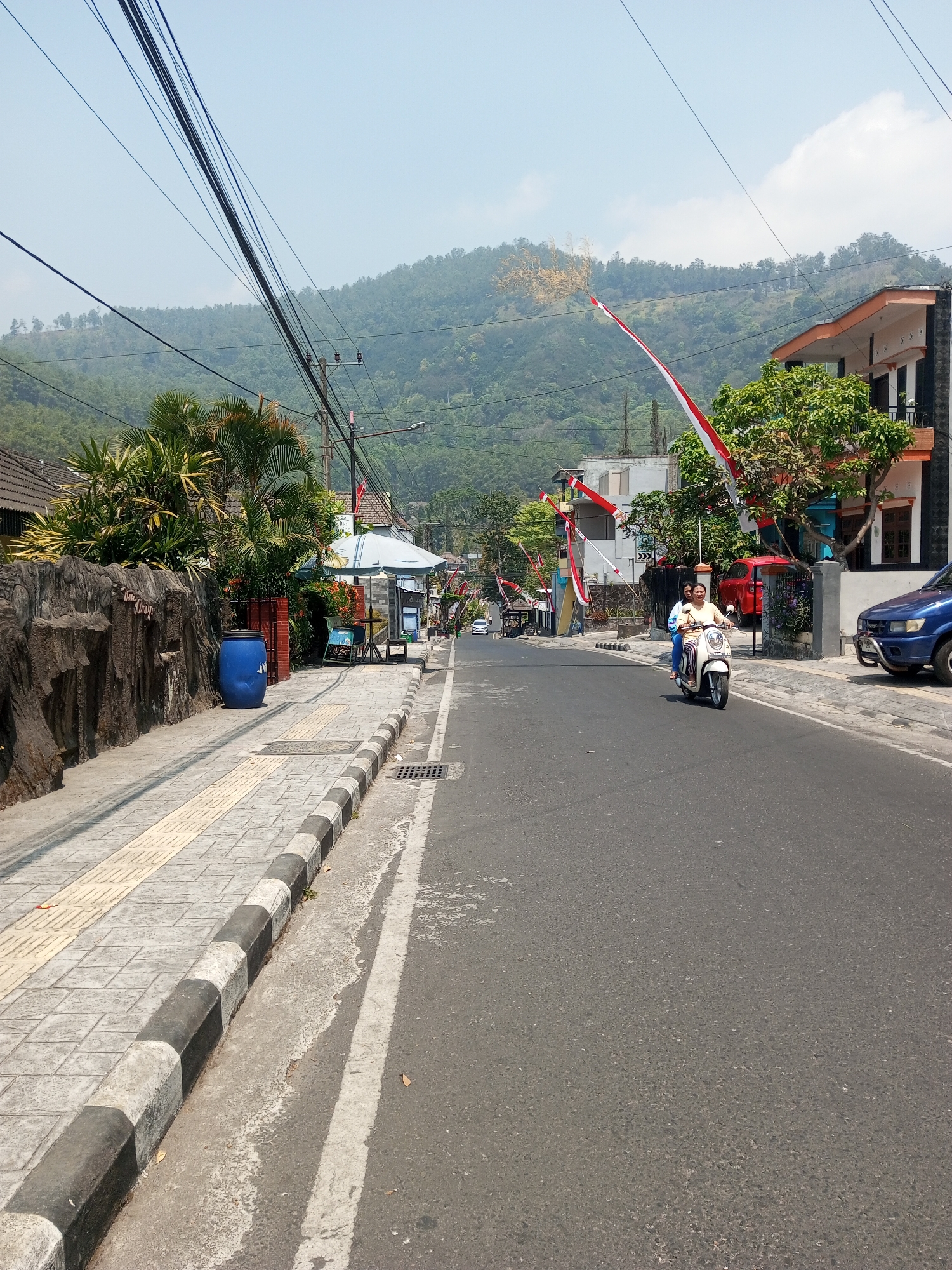 The image depicts a street scene in the city of Batu, Indonesia. A paved road runs through a village-like area, bordered by a sidewalk with a distinctive black and white striped curb. Lush green hills form the backdrop, suggesting a mountainous or hilly terrain. The sky is clear with a few scattered clouds.
On the road, a white scooter is being ridden by two individuals, a woman in the front and a passenger behind her. They are both looking forward, appearing to be traveling down the street. Several red and white Indonesian flags are displayed along the roadside, indicating a possible national celebration or holiday.
Buildings line both sides of the street. On the left, a textured stone wall separates the sidewalk from a property with dense foliage, including palm trees. A large blue barrel sits on the sidewalk near the wall. On the right, a multi-story building with an orange trim is visible, along with a red car parked in front of it and a blue SUV further down the road. Power lines crisscross the sky, supported by utility poles. Small shops or food stalls can be seen further down the street, some with awnings. The overall atmosphere appears to be one of a typical day in a developing urban environment, with signs of local life and national pride.