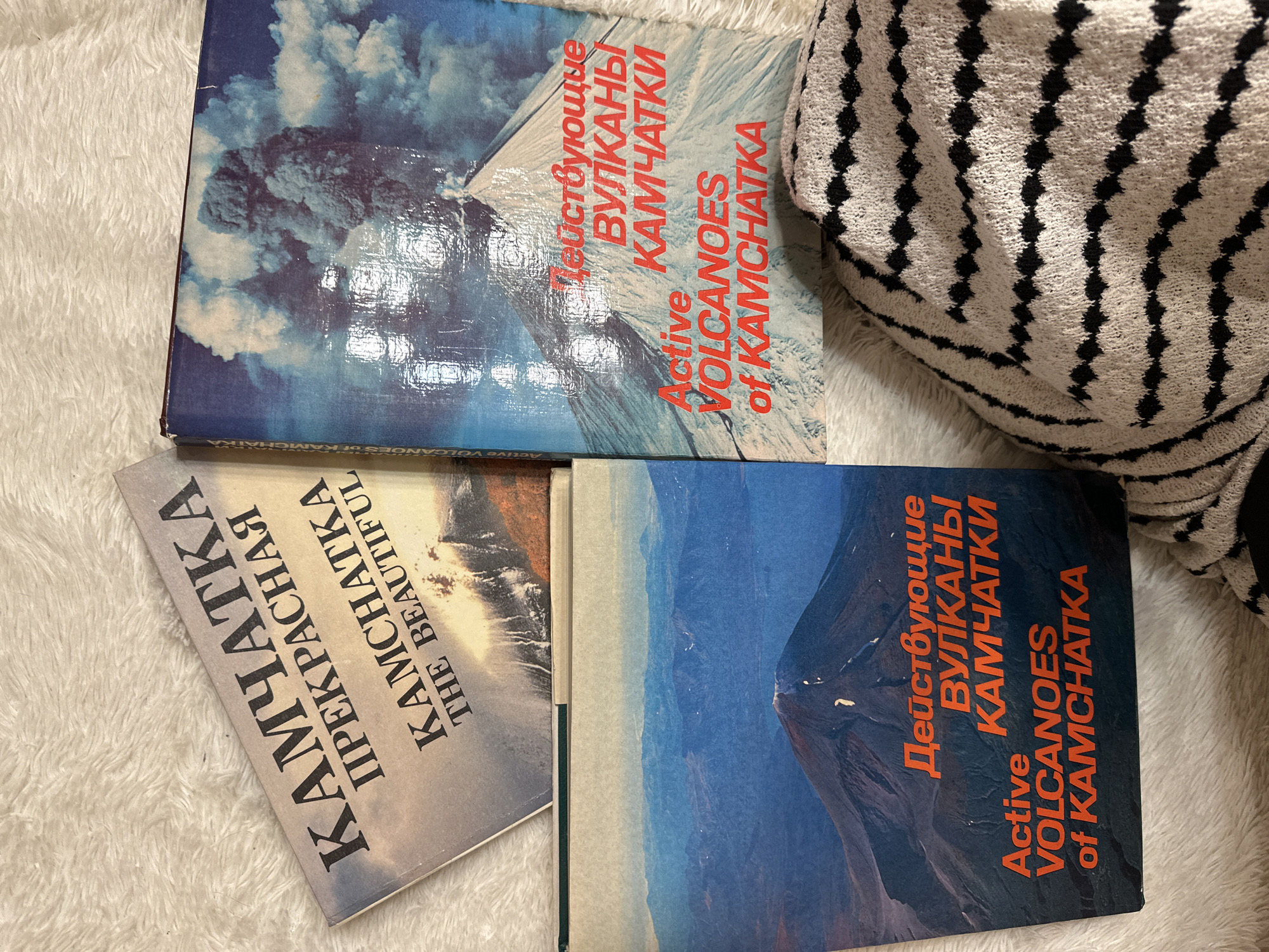 The image shows three books related to Kamchatka, Russia, resting on a textured white surface. Two of the books have the title "Active Volcanoes of Kamchatka" in both English and Russian, with an image of a volcano on their covers. The third book is titled "Kamchatka the Beautiful" in English and Russian, with a landscape image. A patterned black and white item is partially visible in the upper right corner.