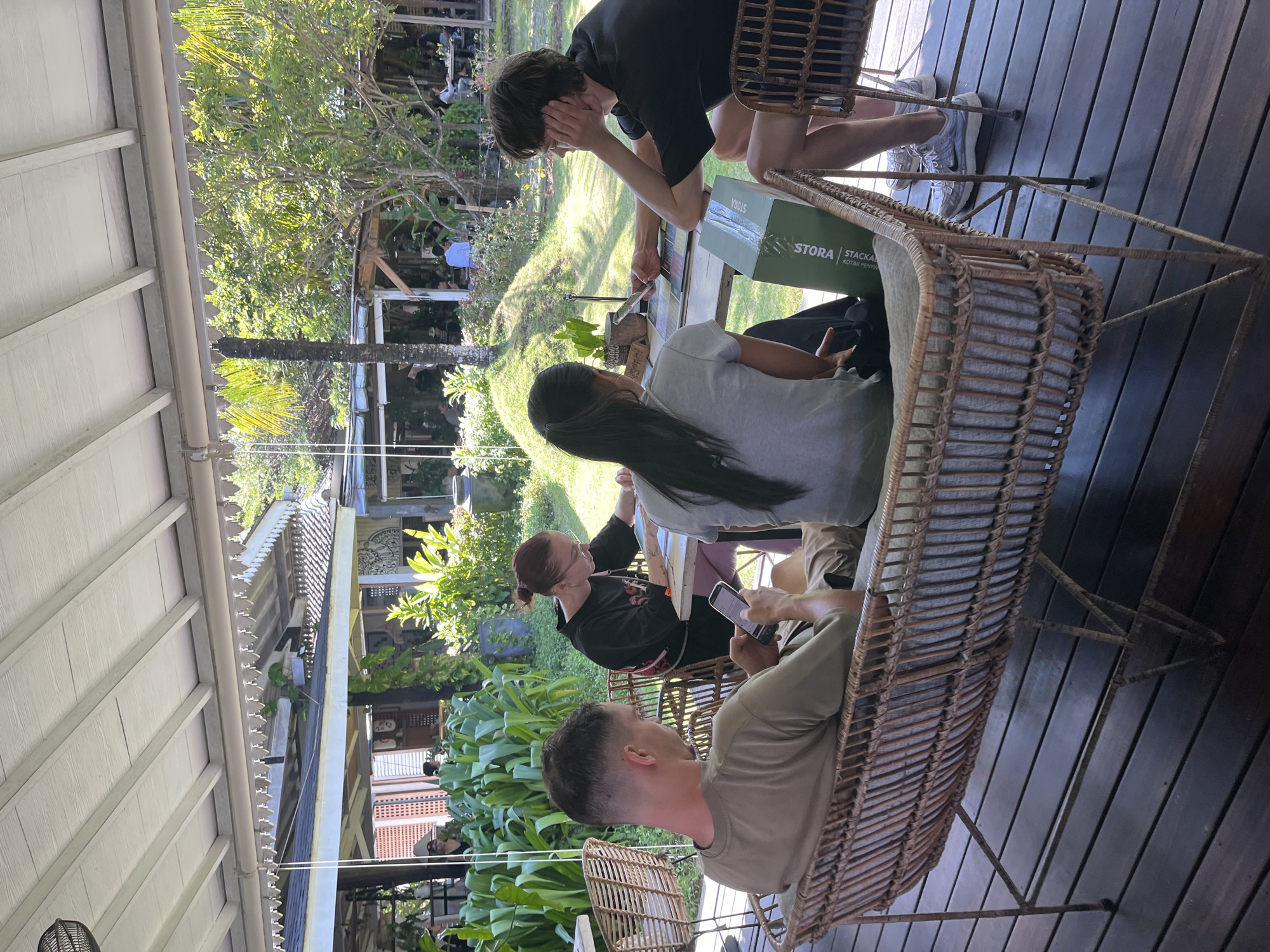 Four people are seated at tables on a covered wooden deck in North Kuta, Indonesia. In the foreground, a man wearing a khaki t-shirt and shorts sits on a wicker bench, looking at his phone. To his right, a woman with reddish hair pulled back in a bun and wearing a black shirt is seated at a table, looking down and appearing to write on a piece of paper. Next to her, a woman with long dark hair in a ponytail, wearing a gray t-shirt, is also seated at the table, looking at her phone. To the right of the women, a young man with dark hair, wearing a black t-shirt and shorts, leans his head on his hand, looking towards the woman in the gray t-shirt. A green box with text "STORA STACKABLE" is on the table in front of him. The deck overlooks a lush, green garden area with several trees and plants. In the background, other people are seated at tables in an open-air restaurant setting. The architecture of the building suggests a tropical, possibly resort-style location.