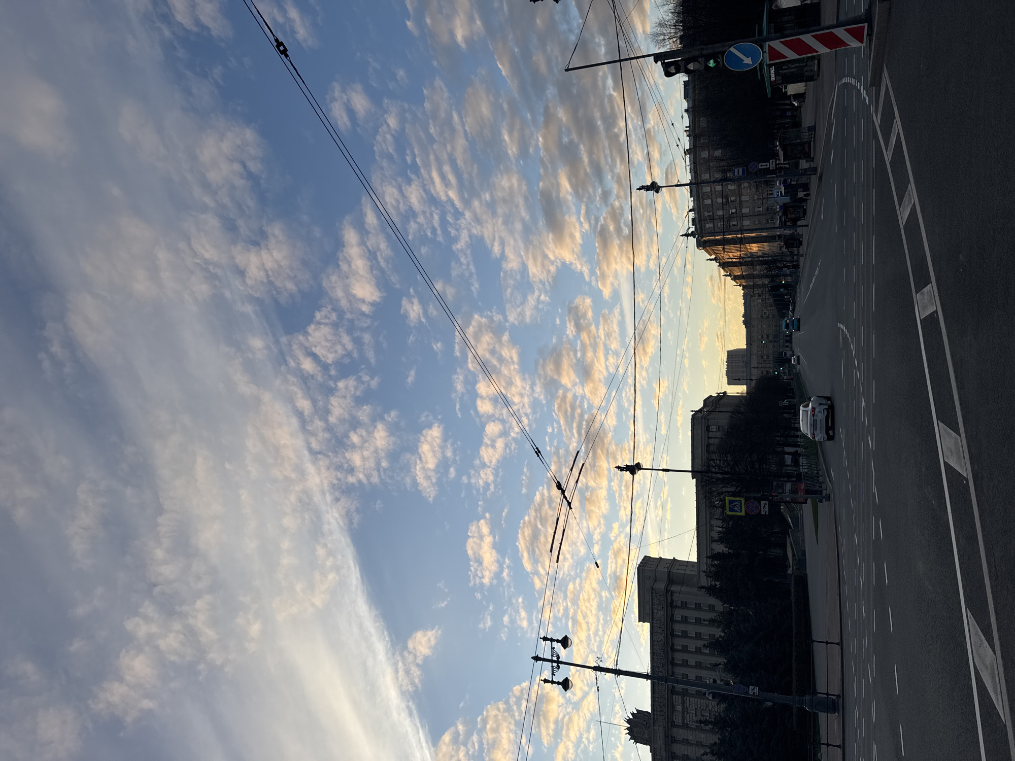 The image captures a street view in Saint Petersburg, Russia. The sky dominates the upper portion of the frame, featuring a mix of blue and white clouds, illuminated by the setting sun. Overhead tram wires cross the sky.
On the right side, the road stretches along the frame, with white dashed lane markings. A white car is visible on the road. Pedestrian crossings and various street signs are visible. Buildings line the street, their architectural details are noticeable, including the distinct Soviet architecture with grand, imposing facades. The buildings' windows reflect the setting sun, adding warmth to the scene. A few trees are silhouetted against the buildings.