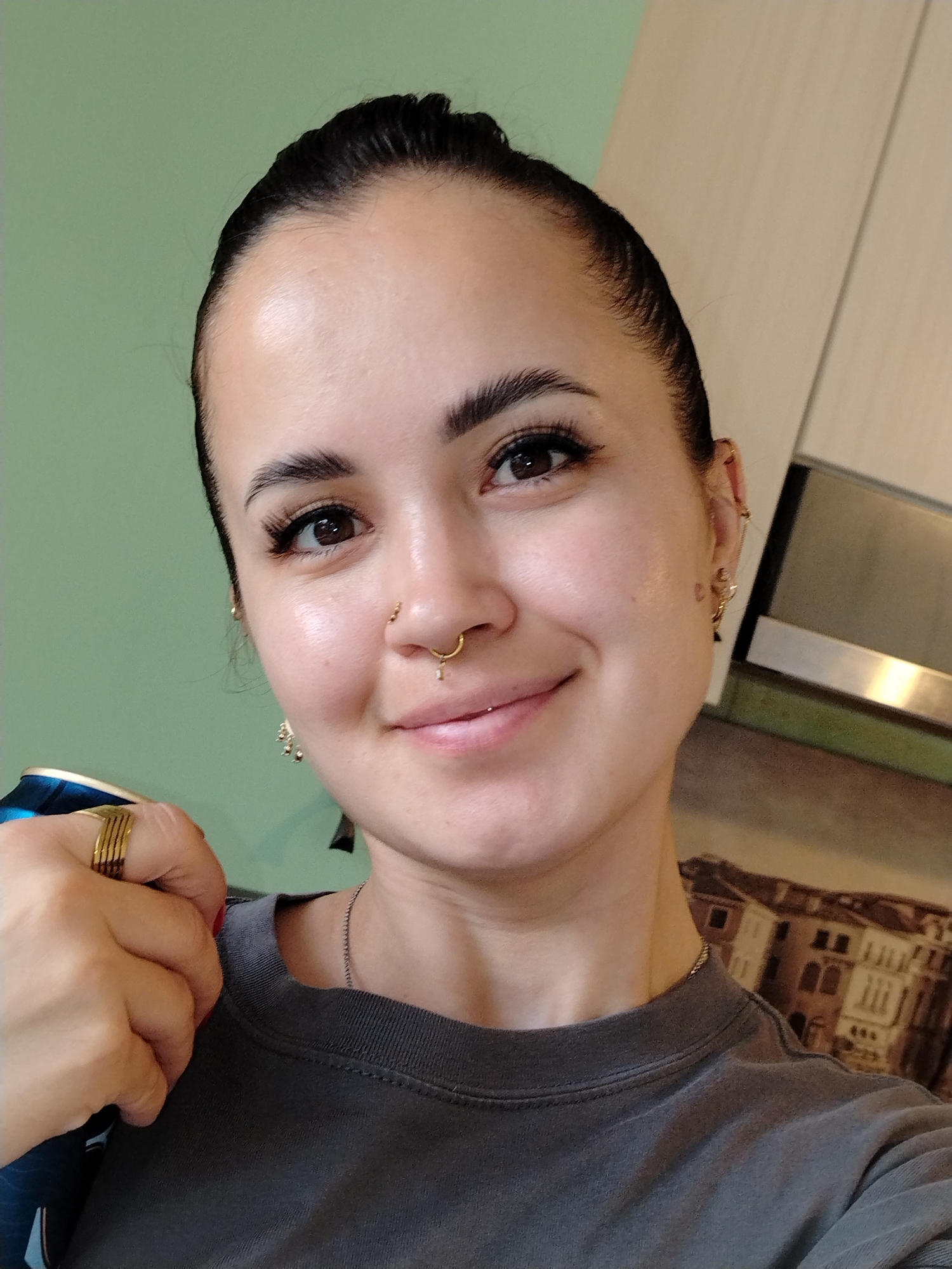 The image shows a close-up, selfie-style shot of a young woman with dark hair pulled back. She is wearing a dark gray t-shirt and has several pieces of gold jewelry, including a ring on her left hand, a septum ring, and earrings. The background is out of focus, but appears to be an indoor setting with light green walls and cabinetry. There is a partial view of a painting or mural in the background depicting buildings. The woman is holding a blue and silver object, possibly a can, with her left hand. She is smiling faintly and looking at the camera. There are no discernible features that would place this image in Noviy, Russia or any other specific city.