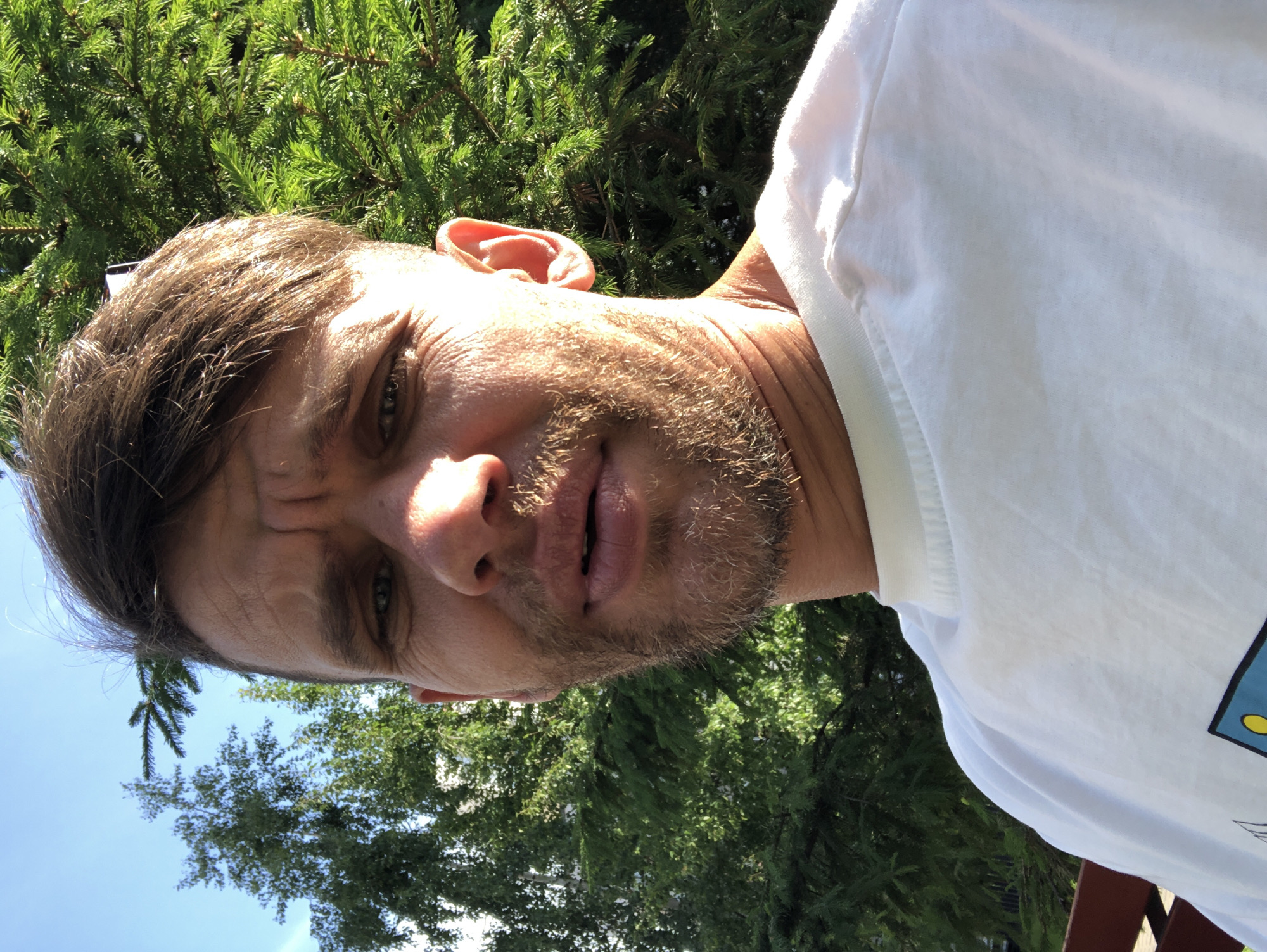 A medium shot selfie shows a white male with brown hair and a light beard looking at the camera. He is wearing a white t-shirt. Behind him, there are green trees and a blue sky. The image appears to have been taken outdoors in natural light. There is no identifiable location or context that would suggest it is in Saint Petersburg, Russia.