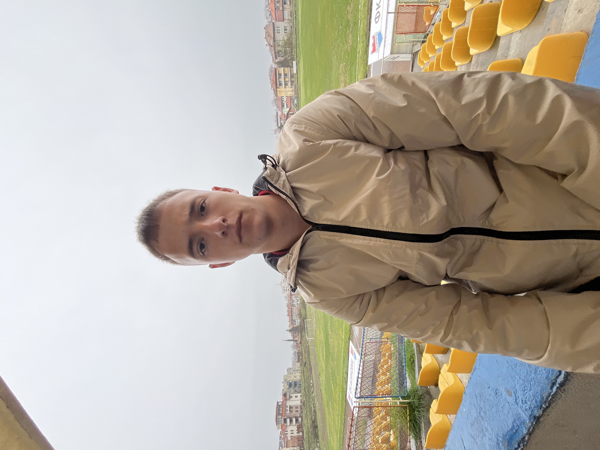 The image is a slightly angled selfie taken by a young man with short blond hair and a fair complexion. He is wearing a light brown jacket with black zippers. The background indicates he is in a stadium, likely in Asenovgrad, Bulgaria, given the cityscape and architectural style visible.
Behind the man, there are rows of yellow stadium seats. A grassy field is visible in the distance. In the far background, there are buildings suggesting a residential area. The weather appears overcast, with a gray sky. A fence or railing is situated behind some of the seats. The stadium is partially visible due to the perspective. The name "Asenovgrad" is displayed on the stadium advertising.