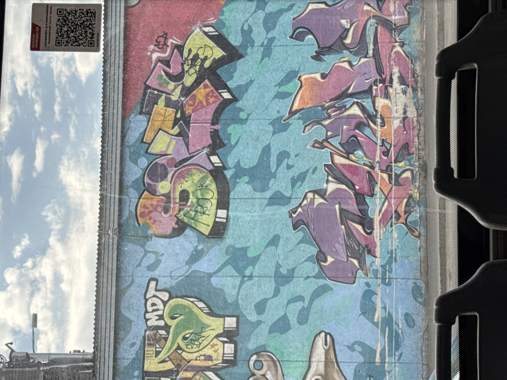 The image shows a colorful graffiti mural visible through the window of public transportation, likely a bus or tram, in the city of Perm, Russia. The mural features abstract shapes, stylized skulls, and cartoonish characters painted in shades of green, yellow, purple, pink, and blue. A QR code related to paying for public transportation is affixed to the window. The sky, with scattered clouds, is seen reflected in the window. Parts of bus seats are visible on the right.