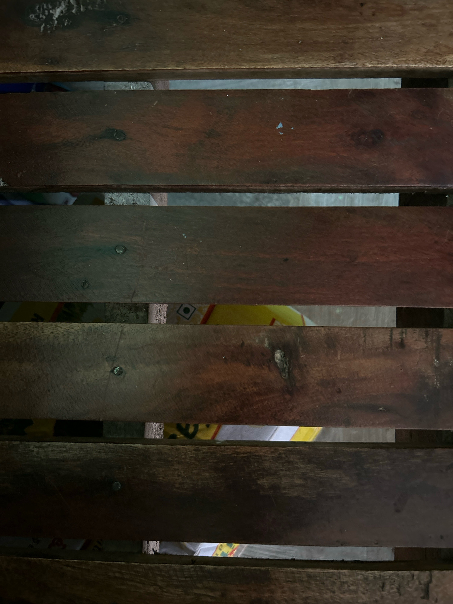 The image shows a close-up, top-down view of several dark brown wooden slats arranged horizontally. The slats appear to be part of a surface, possibly a bench or a pallet, with gaps between them revealing glimpses of what is underneath. The wood grain is visible on the surface, with some areas appearing rougher and more worn than others. There are a few small nails visible, indicating construction. Through the gaps, there are hints of colorful packaging, possibly yellow and white, with some faint geometric patterns. The lighting suggests an outdoor or dimly lit indoor environment. There is no discernible location in the city of Kamareddy, India, or any people or specific actions depicted.