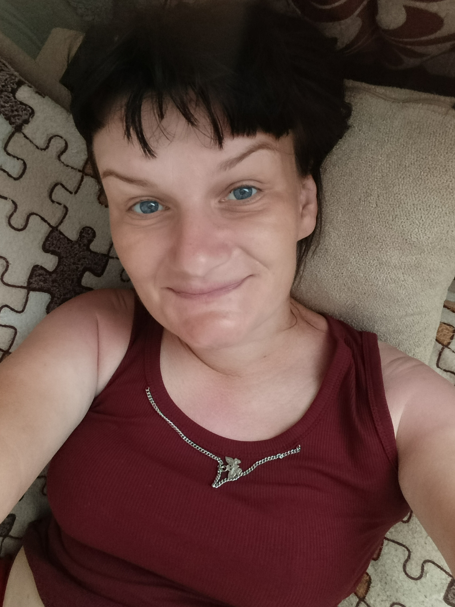 A selfie of a Caucasian woman with short, dark hair and blue eyes is taken from a top-down perspective. She is wearing a maroon ribbed tank top with a silver necklace featuring a small charm. The woman is smiling subtly at the camera. She is lying on a patterned surface resembling a jigsaw puzzle design with beige, brown, and dark brown tones. A beige pillow is positioned near her head. There is no indication that the picture was taken in the city of Фоки, Russia.