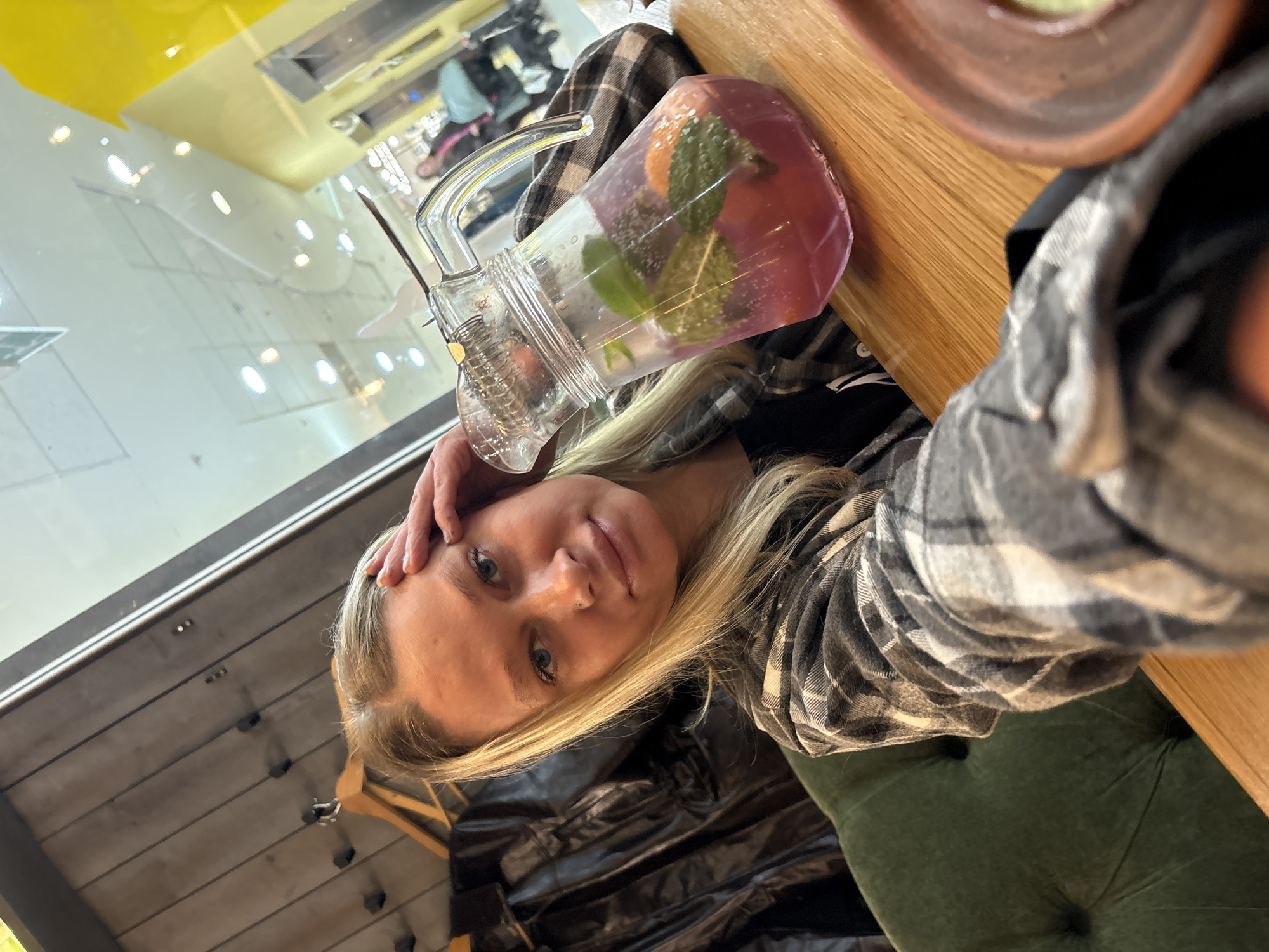 A young, fair-skinned woman with blonde hair is taking a selfie indoors, seemingly at a restaurant or café in Perm, Russia. She is wearing a gray and white plaid shirt. She is holding her hand to her forehead, and her expression is somewhat melancholic.
On the table, there is a glass pitcher filled with a pink-colored drink, likely lemonade, containing mint and orange slices. Next to the pitcher is a brown bowl or plate. The table itself has a light-colored wood finish.
In the background, a window reflects the interior of what appears to be the same establishment, with other patrons and staff visible. The general ambiance suggests a modern, well-lit space. The scene is captured with a slight tilt, adding a casual feel to the image. A dark chair is partially visible at the bottom of the image.