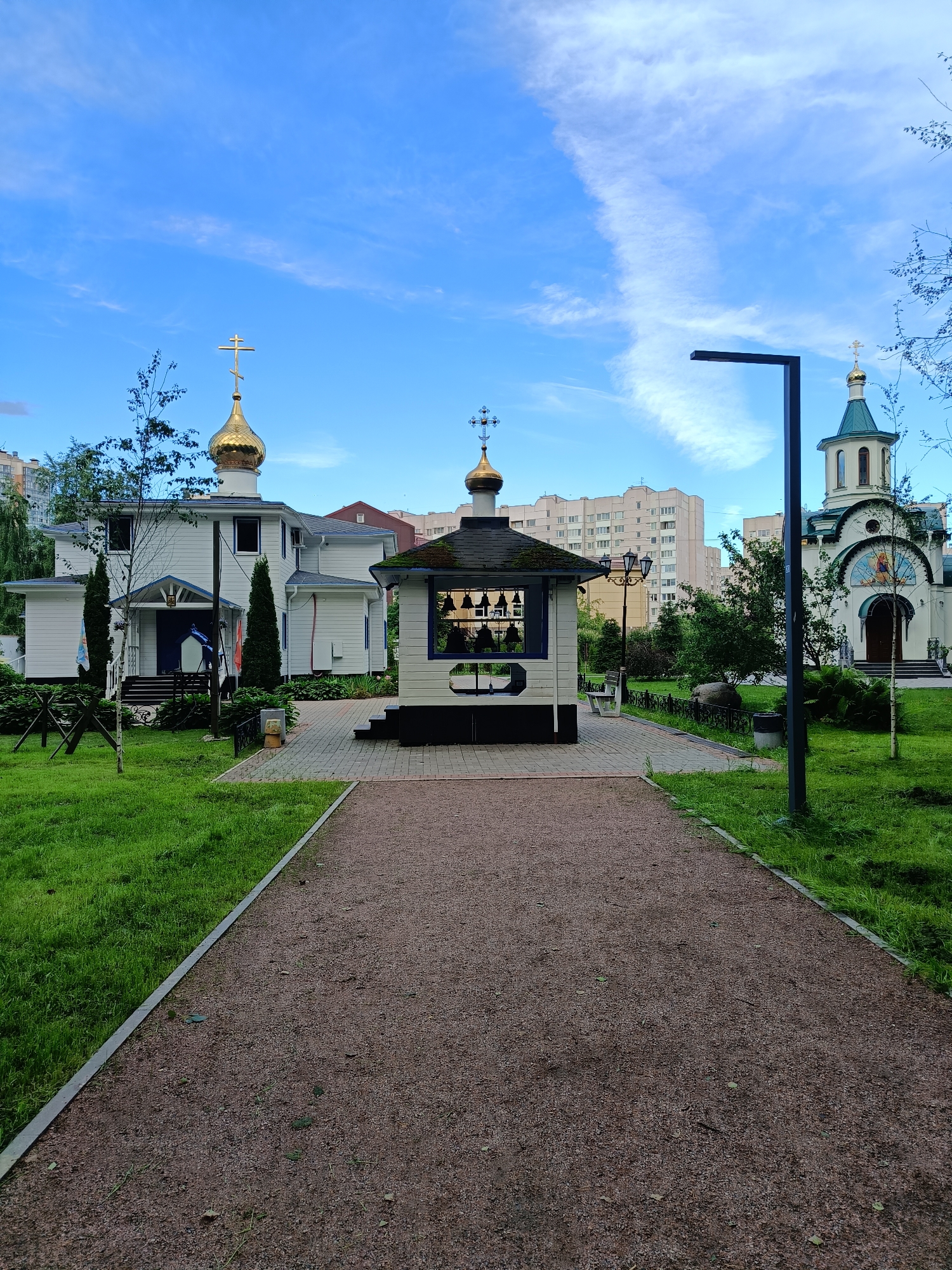 The image depicts a serene courtyard of a church complex in Saint Petersburg, Russia, likely during daylight hours given the bright sky. The foreground features a gravel pathway leading towards the central features. To the left, a lush green lawn is bordered by a paved walkway.
Dominating the scene is a small, white wooden bell tower, octagonal in shape with a dark roof. It houses a set of visible bells. Behind the bell tower, a larger, white wooden church with a golden dome and a Christian cross stands prominently. The church has a visible entrance with steps.
Further in the background, to the right, a second, larger church building is visible. This church has a more traditional design with a green, conical roof topped by a golden dome and cross, and arched windows. Residential buildings, typical of urban St. Petersburg, are visible behind the churches.
The environment is well-maintained, with manicured grass and trees providing a pleasant, natural setting. A modern black lamppost stands on the right side of the pathway, suggesting contemporary urban infrastructure integrated with the religious site. The sky is blue with wispy white clouds. No people are visible in the frame.