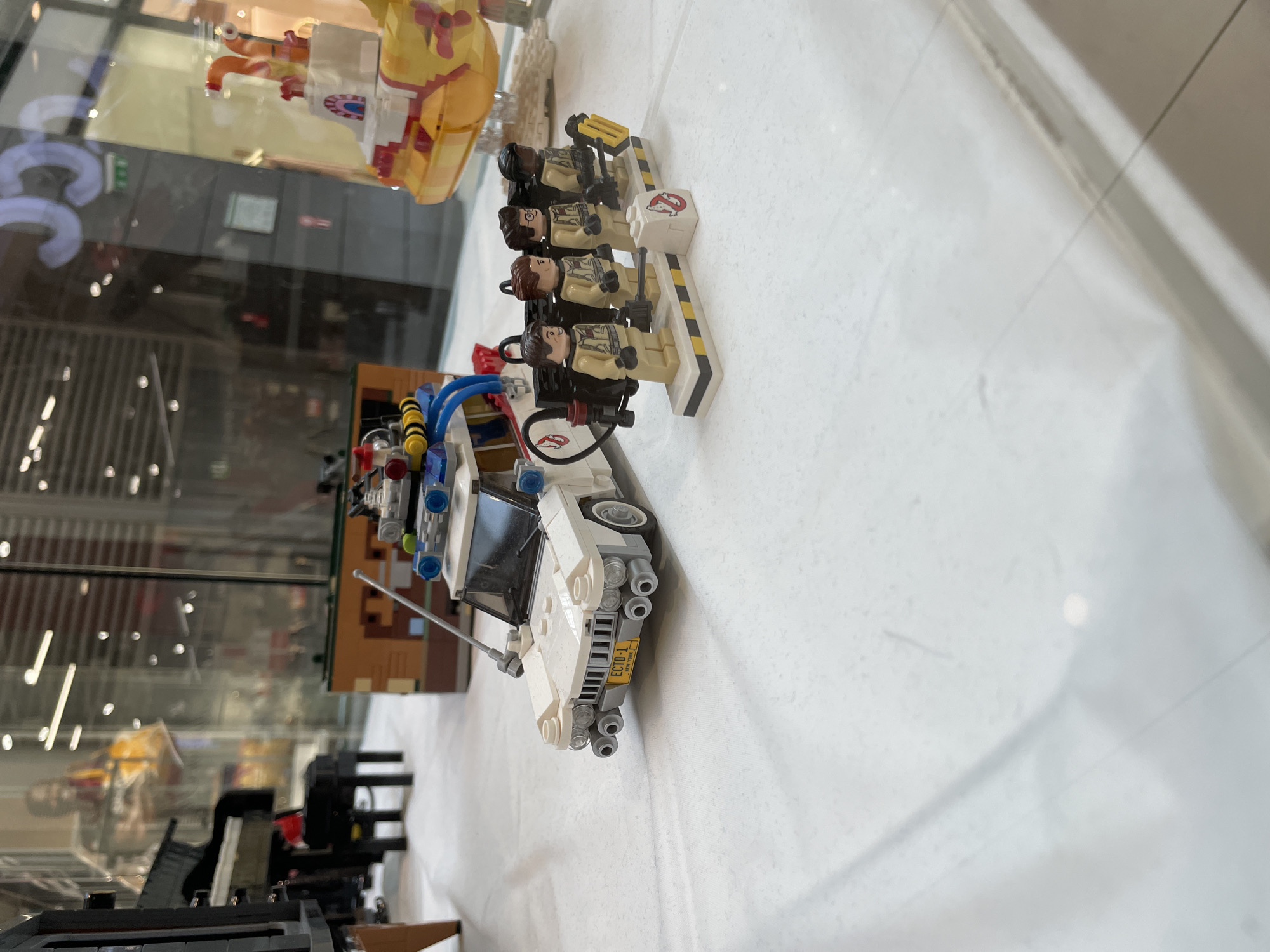 The image shows a display of LEGO models in a glass-covered enclosure. In the foreground, a white LEGO Ecto-1 car from Ghostbusters is parked. Behind the car are four LEGO figures dressed in Ghostbusters uniforms, standing on a platform with proton pack accessories. A small LEGO "No Ghost" sign is also visible. To the left of the car, a section of a LEGO piano and a stool are displayed. In the background, through the glass, there are blurry reflections of store interiors, including signage with the letters "35C" and "55" which may indicate a location within a shopping mall, potentially in Sofia, Bulgaria. A yellow LEGO submarine-like model is partially visible on the right side of the display. The display is set on a white surface, possibly a table or floor, covered with a translucent material.