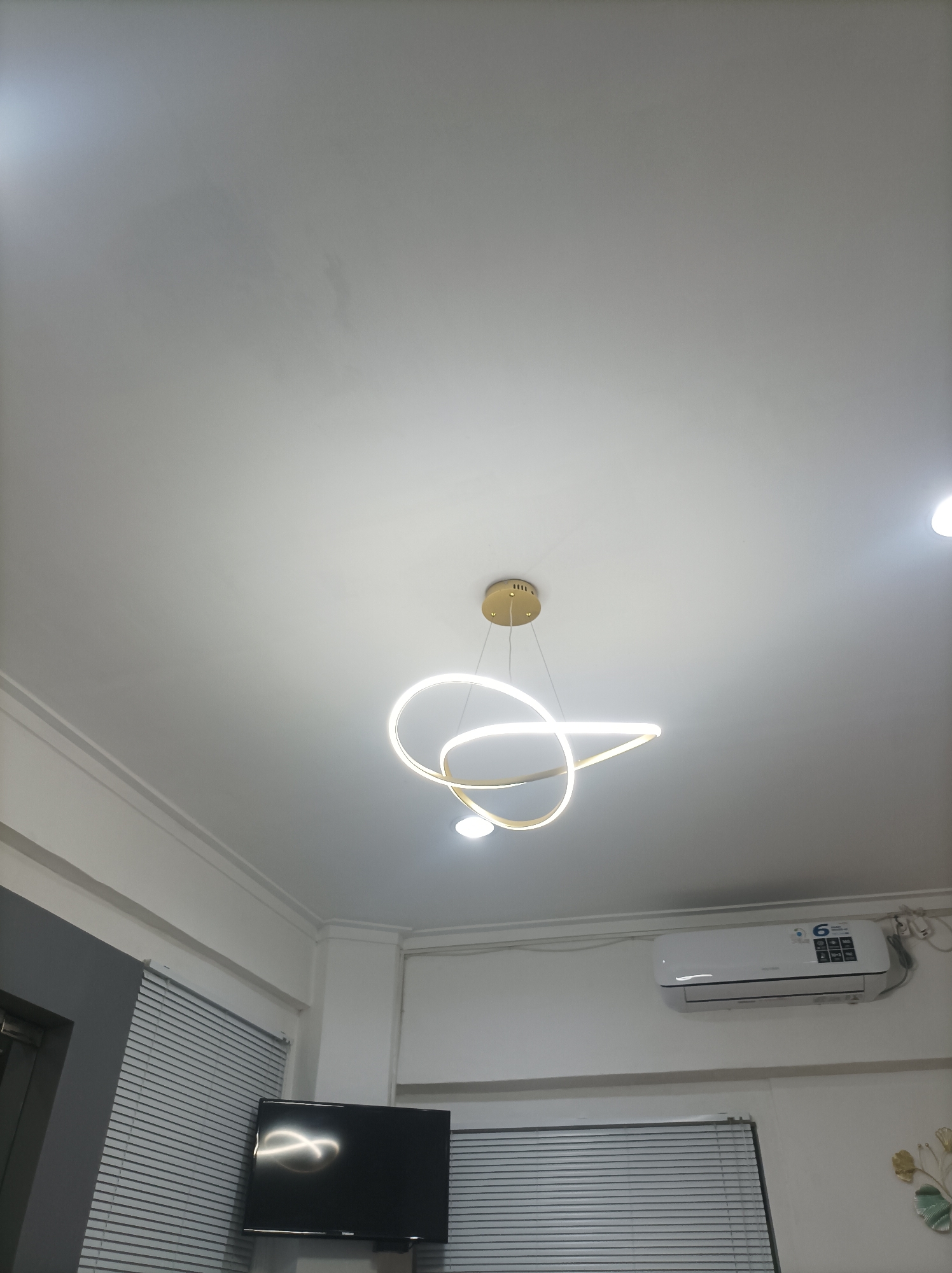 The image shows a modern living room with a white ceiling, a contemporary gold chandelier with two intertwined rings emitting light, and recessed ceiling lights. Below the ceiling, on the wall, is a white air conditioning unit with a digital display. A flat-screen television is mounted on the wall, reflecting the room's lighting. Horizontal blinds cover windows on either side of the television. The room appears to be in a residential setting. The image is captured from a low angle, looking upwards. No people are visible in the image. The overall impression is one of a well-lit and contemporarily furnished interior. The location is identified as Mlilir, Indonesia.
