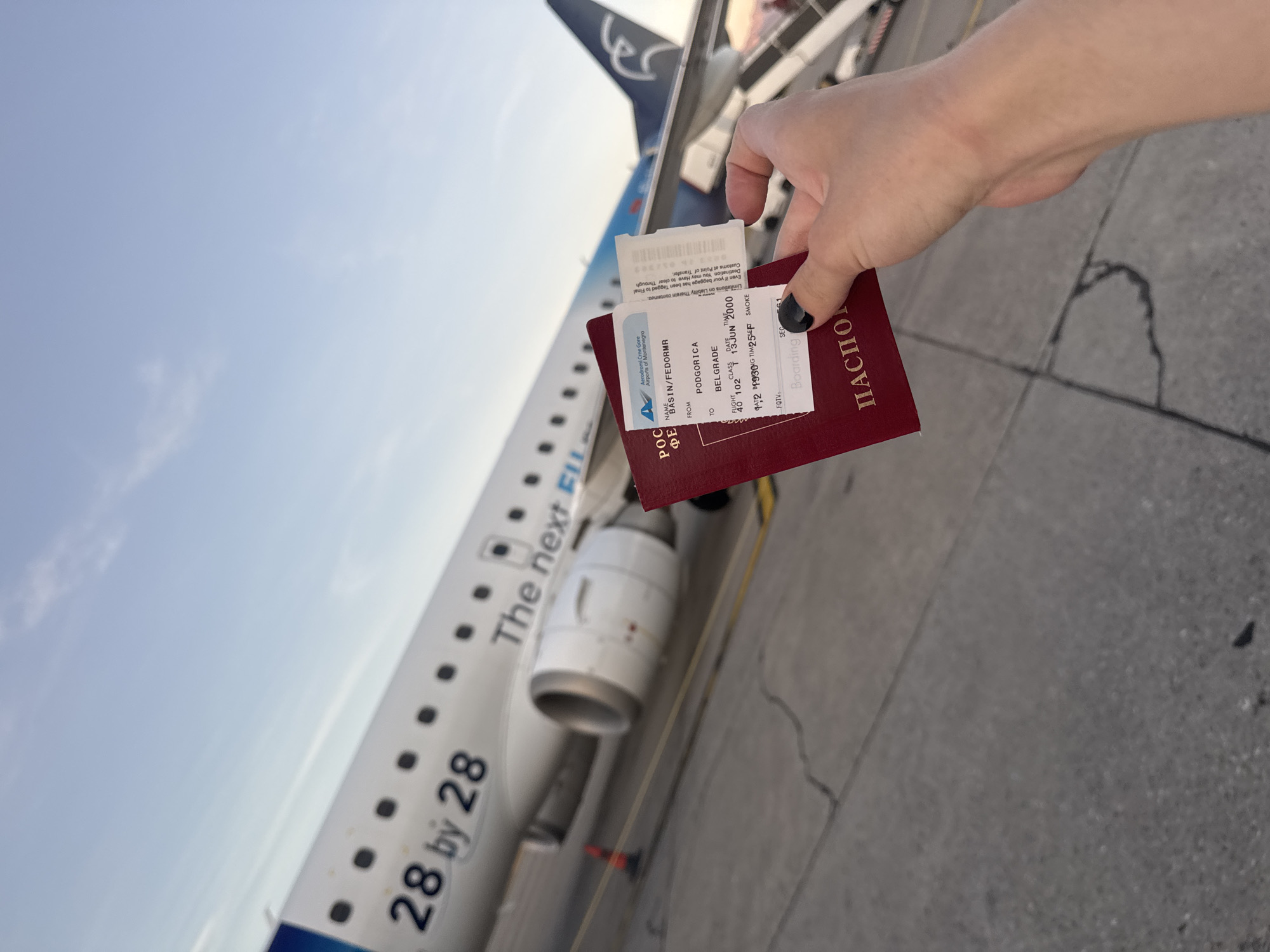 A person's hand holds a red passport and an airplane boarding pass in front of an airplane on an airport tarmac. The boarding pass indicates a flight from Podgorica to Belgrade on June 13, 2000. The airplane appears to be a regional jet with the "28 by 28" logo visible on its side. The sky is clear and blue, suggesting a daytime scene. The location is the Podgorica Airport, which is the international airport of Montenegro.