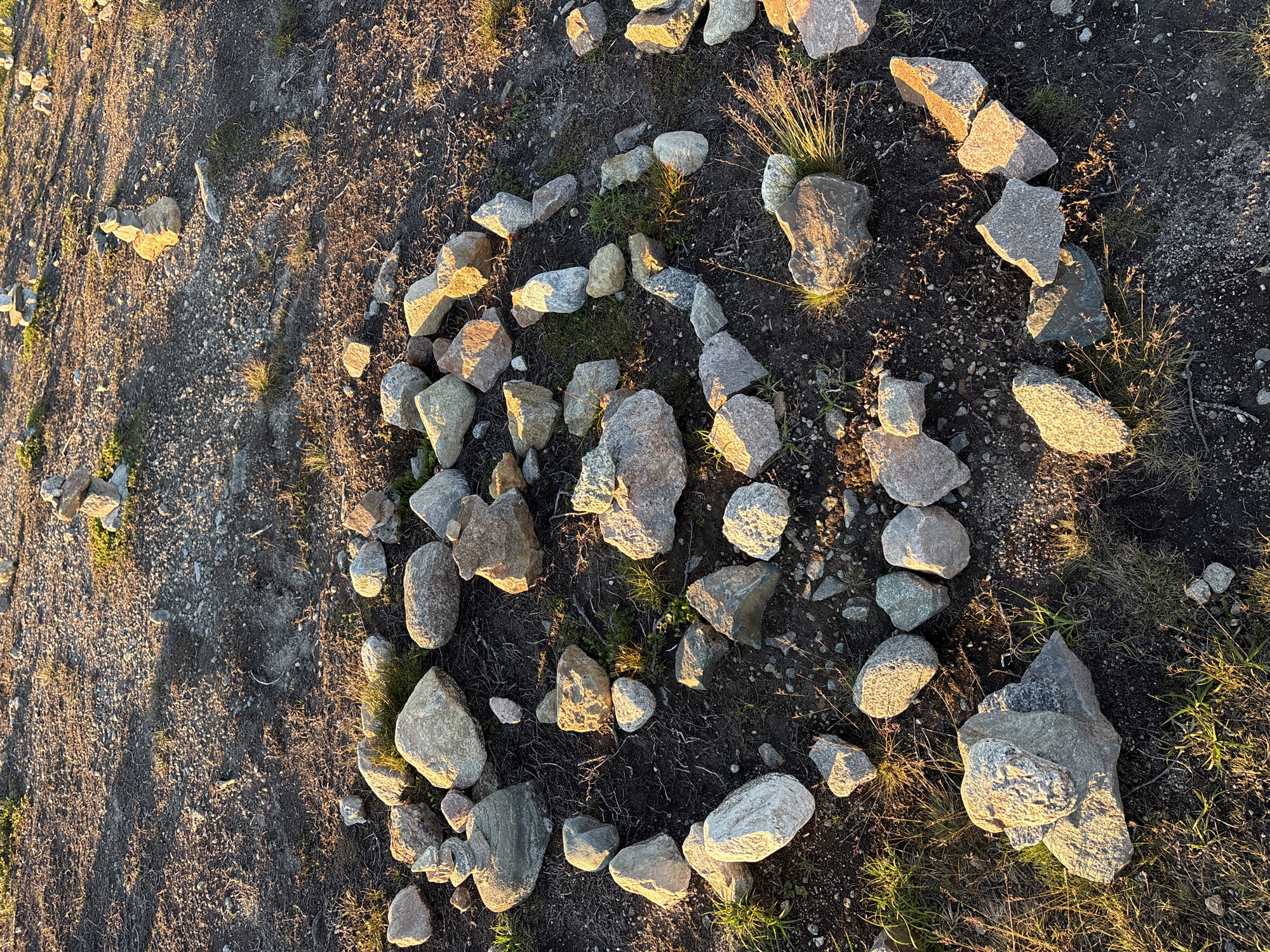 This media file shows an overhead view of a spiral rock formation on the ground, likely outdoors given the presence of dry grass and dirt. The spiral is constructed from numerous grey and tan stones of various sizes and shapes, arranged in a concentric pattern. Some of the stones are smooth and rounded, while others are jagged and angular. The ground surrounding the rock spiral is covered in dry, sparse vegetation and dark soil, with patches of green grass visible. Sunlight casts long shadows across the scene, indicating it is either early morning or late afternoon. The warm, golden light suggests a clear and calm weather condition. There are no people visible in the image, nor any text. The overall impression is one of a natural, perhaps intentionally created, land art installation in a remote or rural setting.