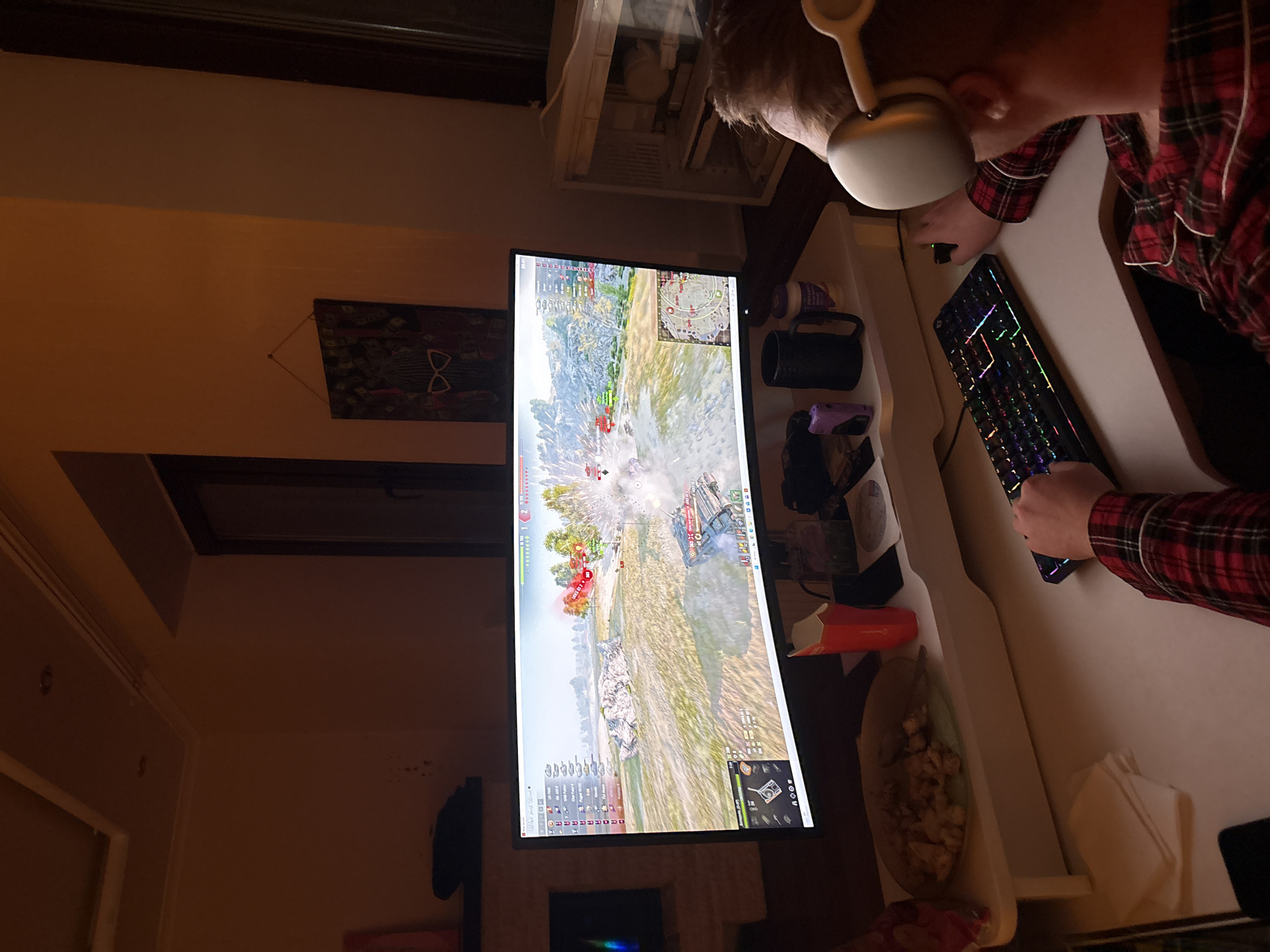 A person wearing headphones and a red and black plaid shirt is playing a tank battle game on a large monitor. A black keyboard with multi-colored lighting is in front of them on a white desk. There is a mug, a purple object, food in a container, and a napkin on the desk. The monitor is displaying the game with a battle scene including tanks, explosions, and terrain. The location is an interior room with a window, wall decoration, and furniture visible. The location in the city of Ореховка, Russia, is unknown.