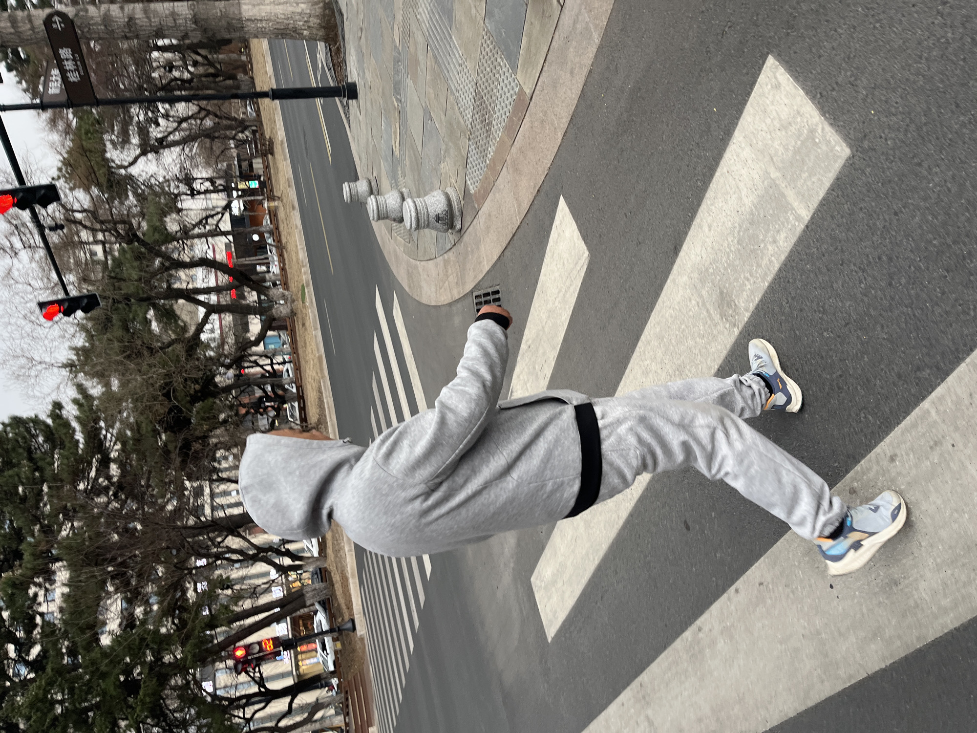 A person wearing a gray hooded sweatshirt and gray sweatpants stands on a crosswalk in Changchun City, China. The crosswalk is marked with white stripes on an asphalt road. To the left, a row of trees lines the street, and traffic lights are visible, showing red signals. A street sign with Chinese characters is mounted on a pole next to the trees. On the right side of the image, a sidewalk with decorative stone elements borders the road. A drain grate is visible near the person's hand.