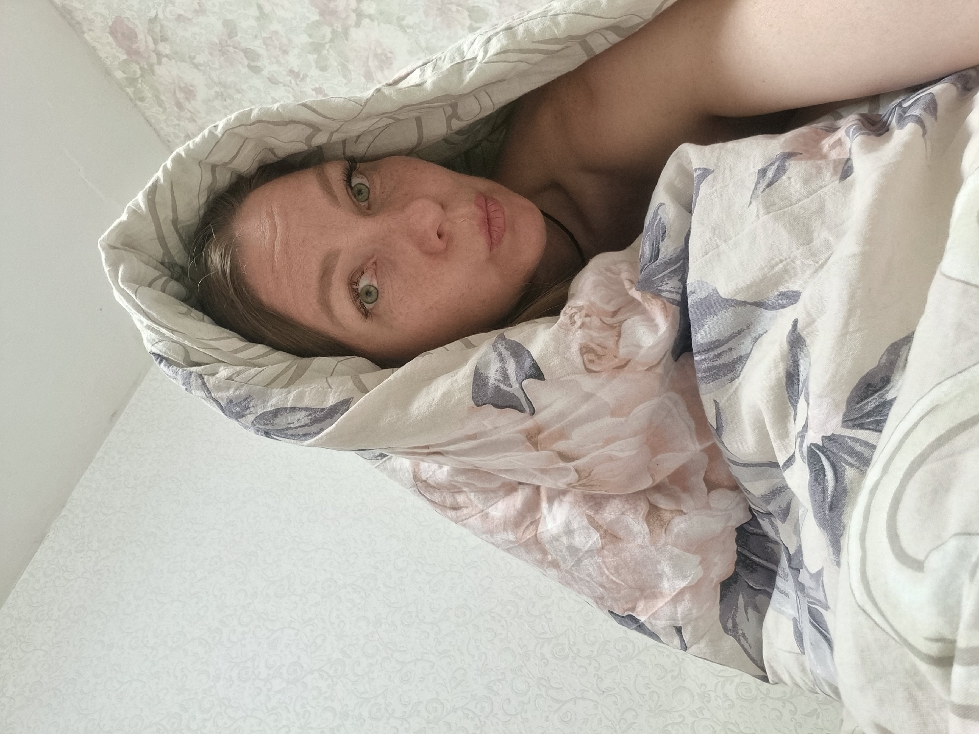 A woman is lying in bed under a floral-patterned comforter. The woman has light skin, green eyes, and blonde hair. She is looking directly at the camera with a neutral expression. The comforter is pulled up around her head, creating a sort of cocoon effect. The wallpaper in the background is light-colored with a subtle floral pattern. The image was likely taken indoors in a residential setting, possibly in the city of Chaikovsky, Russia.