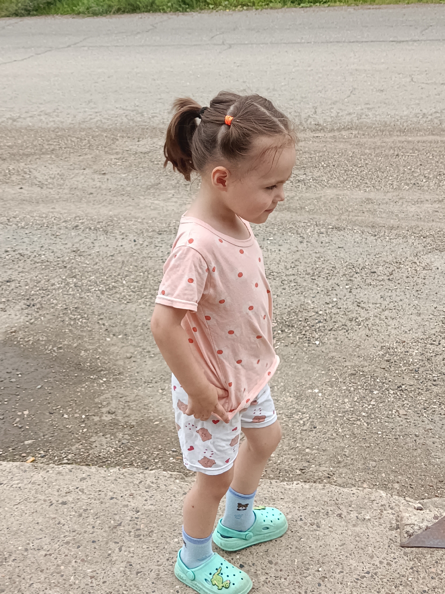 A young girl with brown hair in two ponytails walks on a concrete path next to a gravel road. She is wearing a light pink t-shirt with red polka dots, white shorts with a pattern of bears and hearts, light blue socks with a bear design, and light green clogs. She is looking down and to the right. The background is a gravel road with some green foliage in the distance. The scene appears to be outdoors, possibly in a residential area or a park, in the city of Posad, Russia.