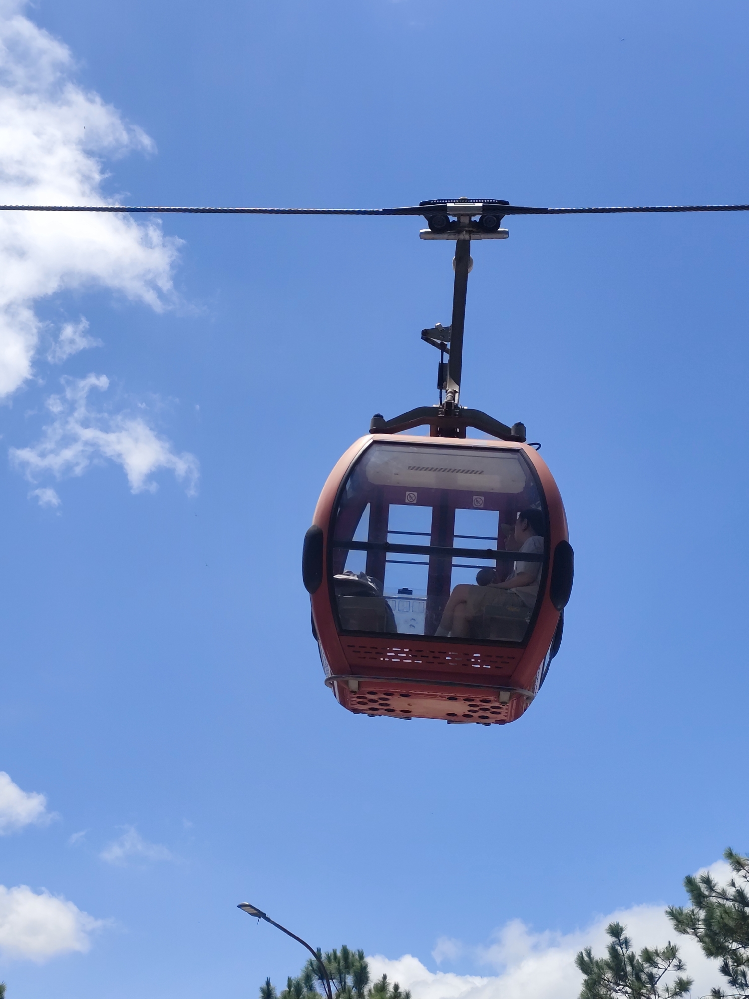 The image shows a red cable car suspended in the air against a bright blue sky with scattered white clouds. The cable car has large windows, and a person can be seen sitting inside. In the lower part of the image, there are green pine trees and a lamppost. This is likely the cable car system in Da Lat, Vietnam, which offers scenic views of the city and its surroundings, including the Tuyen Lam Lake and the Robin Hill.