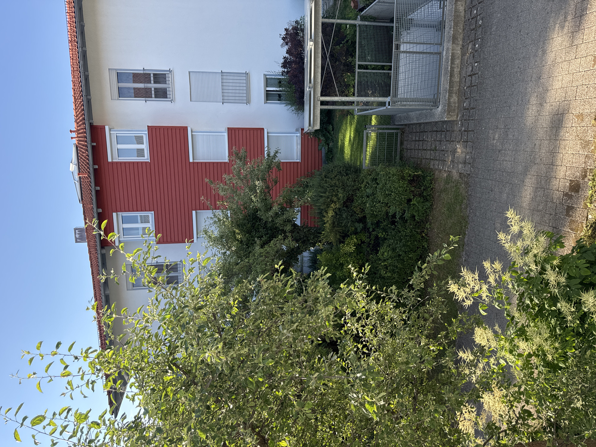 The image shows the exterior of a multi-story building with a red and white facade, located in Höhenkirchen-Siegertsbrunn, Germany. The building features several windows with roller shutters and a small balcony. In front of the building, there is a garden area with lush green trees and bushes, some of which are in bloom. A paved walkway leads to a covered structure with metal fencing, possibly an entrance to a basement or storage area. The sky is clear and blue, indicating a sunny day. The overall scene is residential and peaceful.
