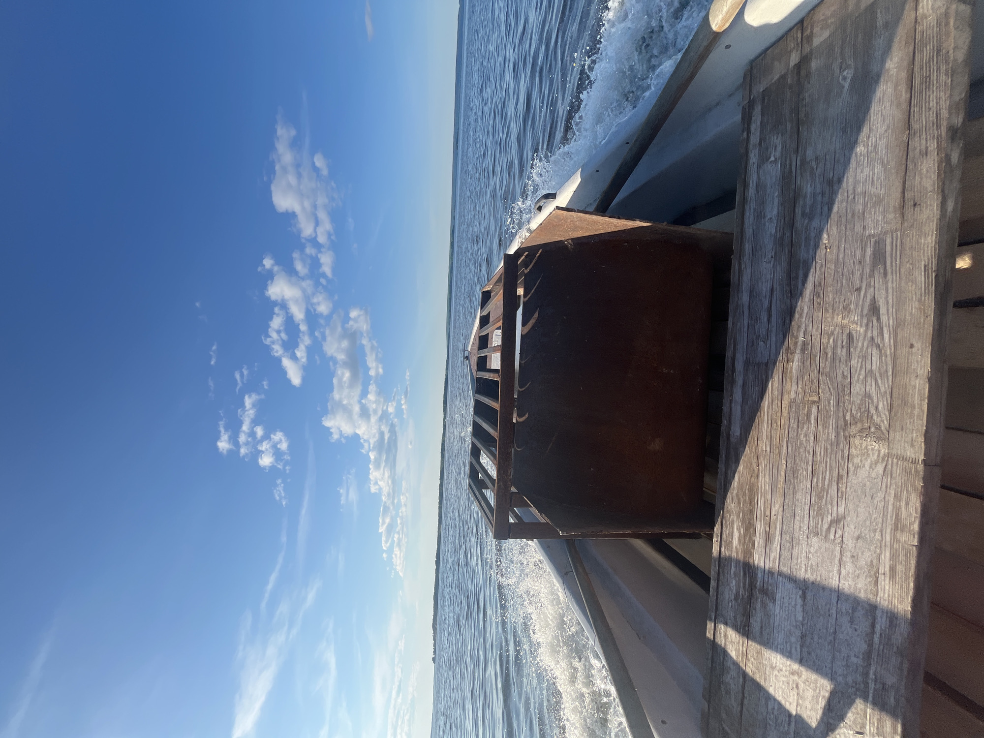 The image shows a view from the back of a boat moving on a body of water. The boat's interior is visible in the foreground, with weathered wooden planks forming part of the seating or deck. A large, rusty metal object, possibly a cage or container, is situated in the center of the boat. White wakes are trailing from the boat, indicating forward motion. The water is calm with small ripples, and a distant shoreline with trees is visible under a clear blue sky dotted with scattered white clouds. The lighting suggests it is daytime, likely late afternoon or early morning due to the low angle of shadows cast on the wooden planks. The scene appears to be in a rural or natural setting, consistent with the location of Vurmankasy (Vurman-Siukterskogo S/P), Russia.