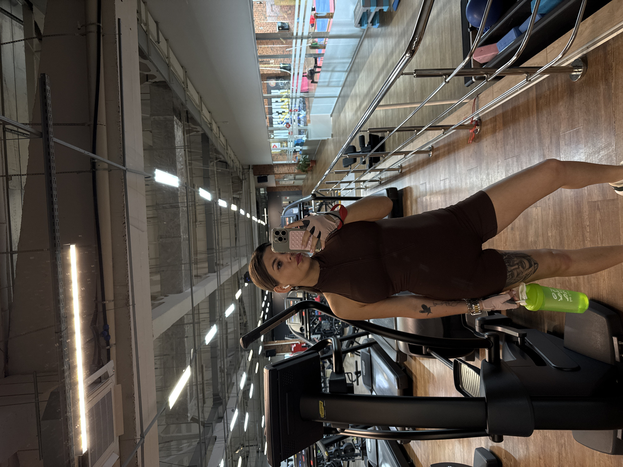 The image shows a young woman with dark hair pulled back, taking a mirror selfie in a brightly lit gym. She is wearing a brown sleeveless leotard and white athletic gloves. She is holding a bright green shaker bottle in her left hand. The gym has wooden floors, a row of treadmills on the left, and various weight machines and exercise equipment visible in the background. A metal railing runs along the right side of the image. The ceiling features exposed industrial lighting fixtures and ductwork. The overall impression is of a modern fitness facility. The image does not contain any specific visual cues to indicate it is located in Chaikovsky, Russia.