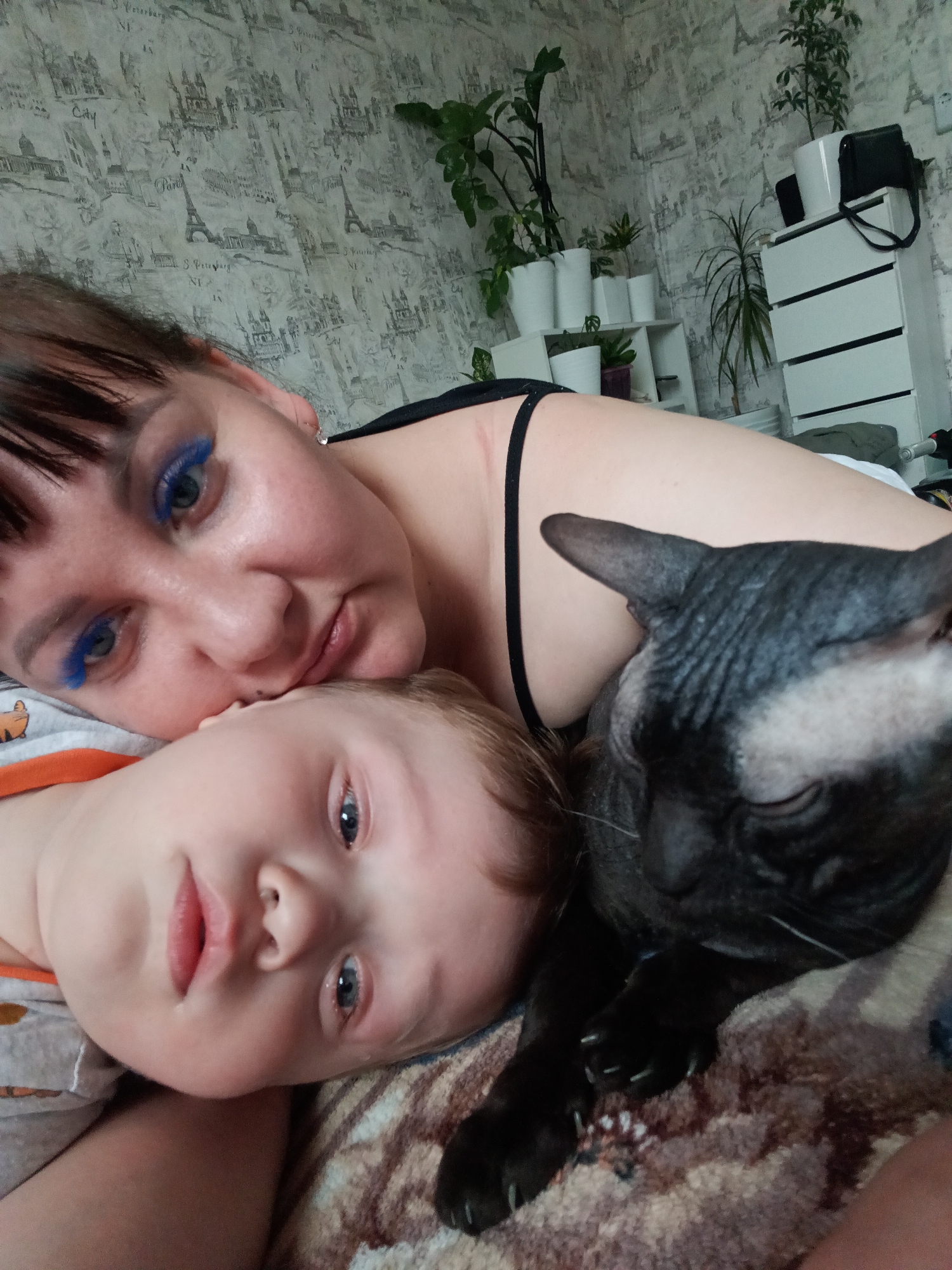 The image is a selfie taken indoors, featuring a woman, a child, and a black cat lying on what appears to be a bed or couch. The woman has dark hair with bangs and is wearing bright blue eyeshadow. The child, with light-colored hair and eyes, is positioned beneath her. A hairless black cat is positioned beside the child. The background includes wallpaper with a pattern of cityscapes, including depictions of Paris and St. Petersburg, as well as potted plants on a shelf and a white dresser. The location of Osa, Russia, is not identifiable from the image.