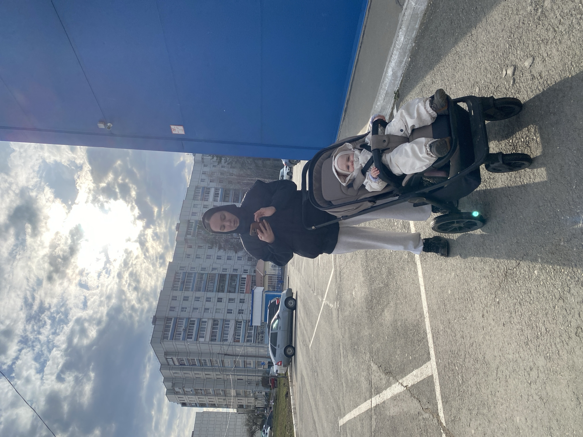 An outdoor image captures a woman pushing a baby in a stroller across a paved parking lot in Chaikovsky, Russia, on a partly cloudy day. The sun is bright, breaking through the clouds in the upper left of the frame, casting distinct shadows.
The woman, with dark hair, wears a black hooded jacket, light-colored sweatpants, and dark sneakers. She holds a smartphone in her right hand, looking down at it. Beside her, a baby is seated comfortably in a dark-colored stroller, dressed in a warm, light-colored padded outfit with a matching hooded hat. The baby looks towards the camera with a calm expression. The stroller has visible wheels and a safety harness.
In the background, a large, tall blue building or structure dominates the left side of the scene. On its facade, a small, red rectangular sign with indecipherable white text or symbols is visible, along with a security camera mounted higher up. Beyond the blue structure and across the parking lot, several multi-story residential apartment buildings with numerous windows are visible. A light-colored sedan is parked in the lot. The ground is asphalt with white parking lines, some of which are faded. The sky is filled with both bright light and scattered clouds.