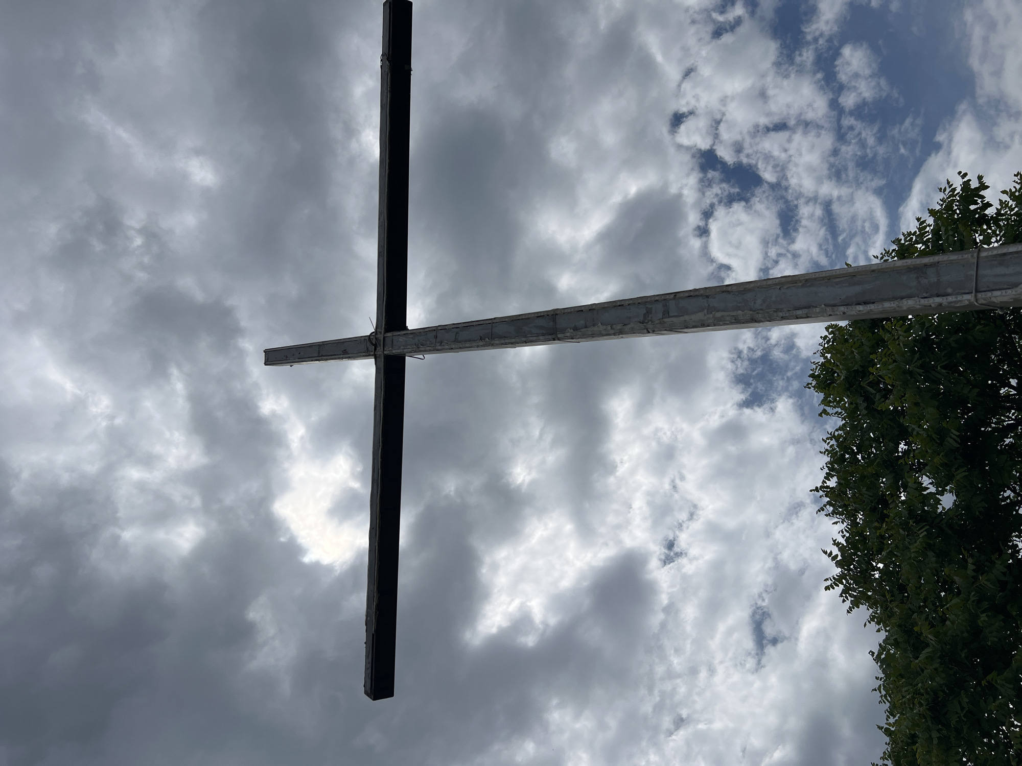 A low-angle shot shows a metal cross against a cloudy sky. The vertical beam of the cross is dark and solid. The horizontal beam is a lighter grey, seemingly constructed of multiple pieces connected by wires. Parts of the sky are light blue with white clouds, while other parts are covered in dense, grey clouds. A portion of a tree with green leaves is visible in the bottom right corner. This is possibly the metal cross located in Asenovgrad, Bulgaria.
