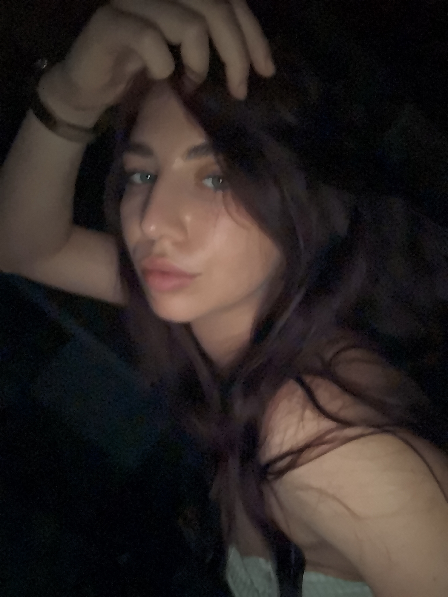 A close-up photograph shows a young woman with long, dark hair. She is looking directly at the camera with a relaxed expression. Her left hand is raised to her forehead, obscuring part of her hair. She is wearing a bracelet on her left wrist. The background is dark and indistinct, suggesting a dimly lit interior. Location: Unknown, Unknown.