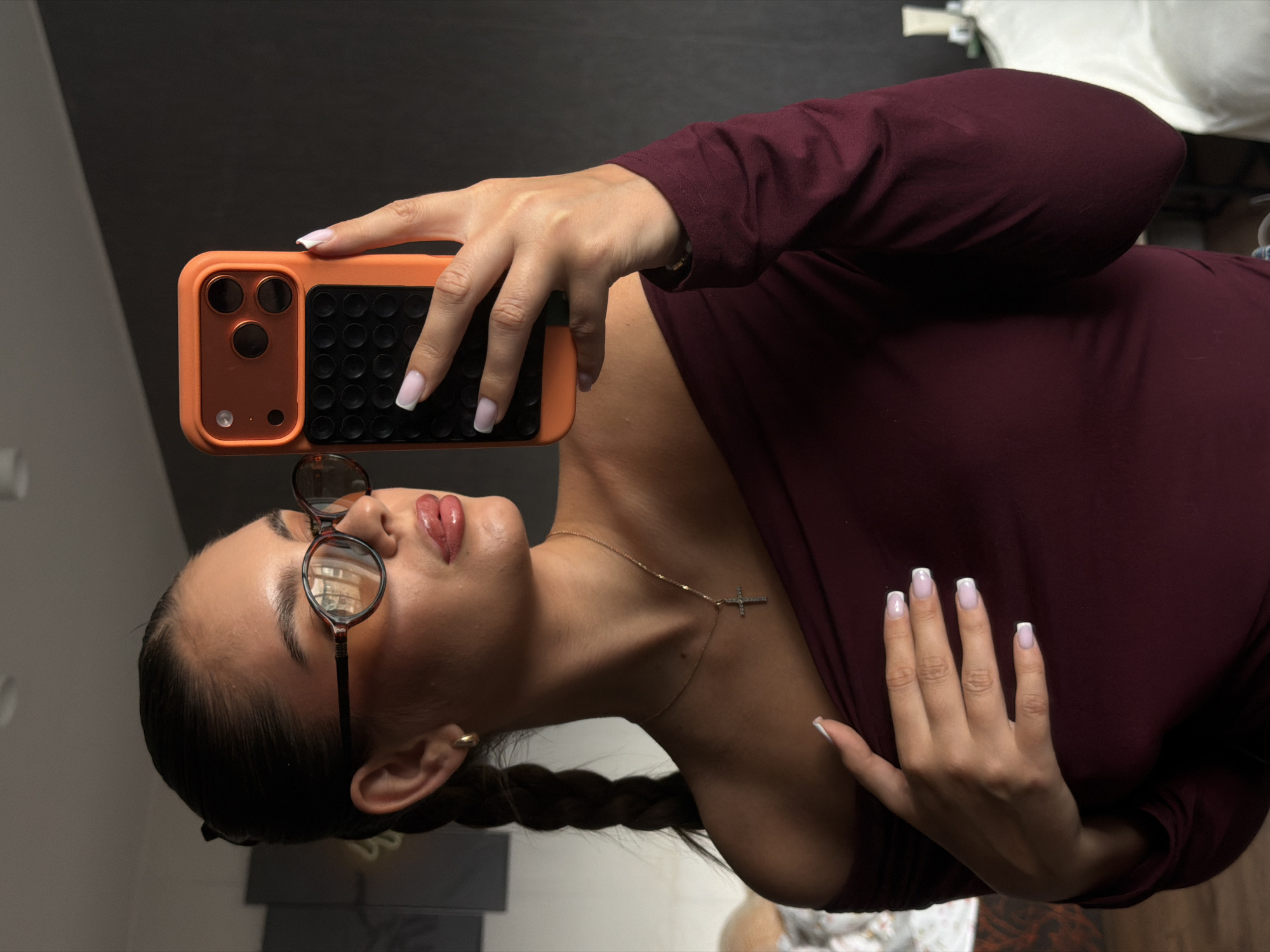 A young adult female with medium brown hair braided down her back and wearing round-framed glasses is taking a selfie. She is holding an orange phone case in her left hand. Her nails are manicured with a white tip. She is wearing a dark burgundy long-sleeved top. A gold necklace with a cross pendant is visible around her neck. The background appears to be a modern interior with a dark gray surface. It is impossible to determine the location as Krasnoyarsk, Russia, based on this image alone.