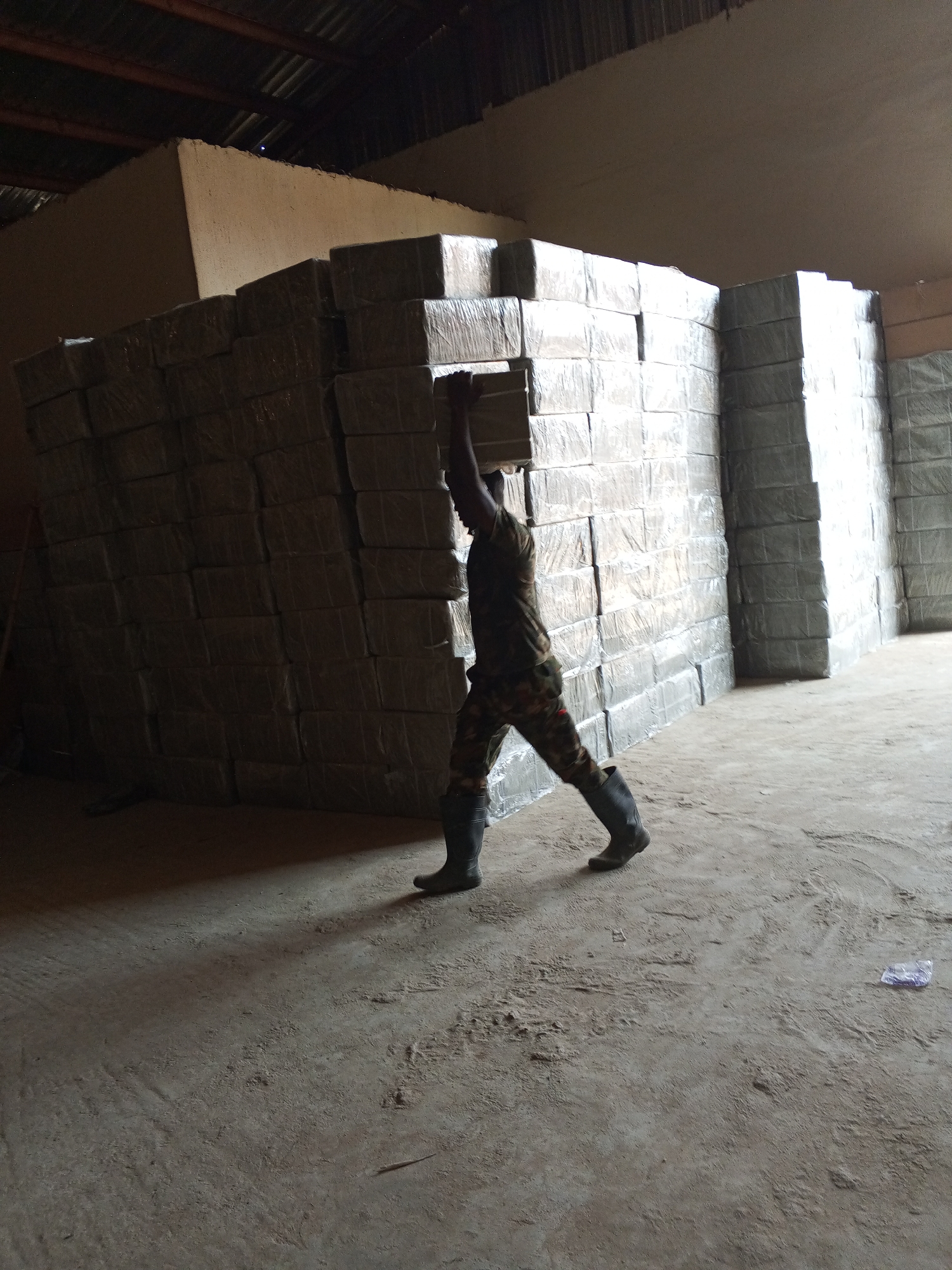 A man wearing camouflage clothing and black rubber boots is carrying a rectangular, packaged object on his head in a large warehouse. Stacks of similar objects, wrapped in a transparent film, line the walls of the warehouse. The floor is concrete and slightly dusty. The location is presumed to be in Pasere, Nigeria, based on external information.