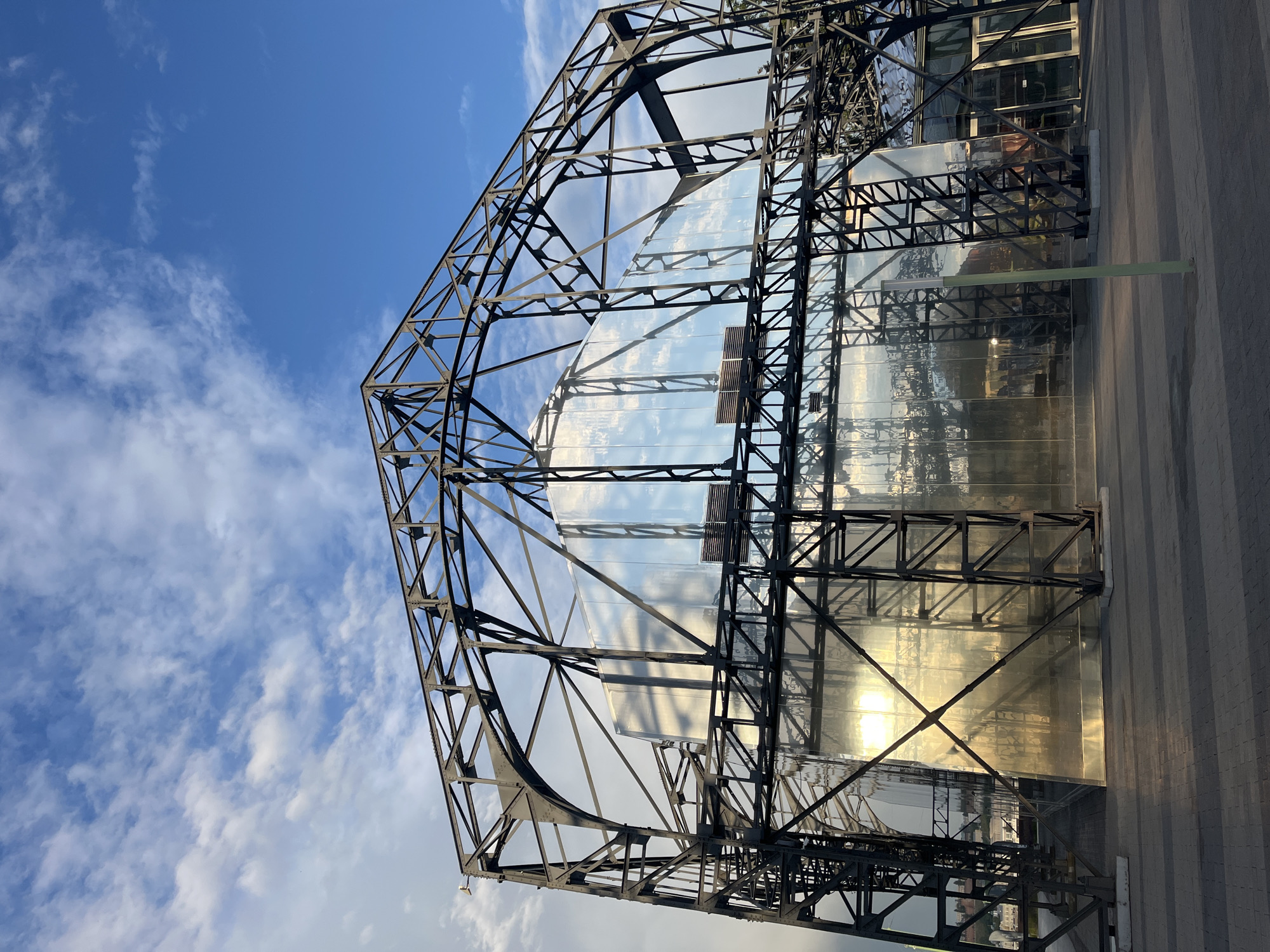 The image shows a low-angle view of a large, metallic, geodesic-like structure with a clear blue sky and scattered clouds above. The structure appears to be a building with a complex framework of dark metal beams. Large, reflective glass panels are visible within the structure, mirroring the sky and surrounding environment, including what seems to be other buildings and trees. Sunlight glints off the glass, indicating it is daytime.
To the right of the structure, a paved area with a repeating pattern of rectangular stones is visible. A thin, light green rectangular object extends horizontally from the structure over this paved area. The overall scene suggests an outdoor urban setting, possibly a park or a public square, in Nizhny Novgorod, Russia. The geometric nature of the structure and the reflective surfaces create a modern and potentially architectural display. No people are visible in the image. The time of day appears to be late afternoon or early evening, given the angle of the sunlight and the long shadows cast on the paved area. The weather is clear with some white, wispy clouds. There is no visible text in the image.