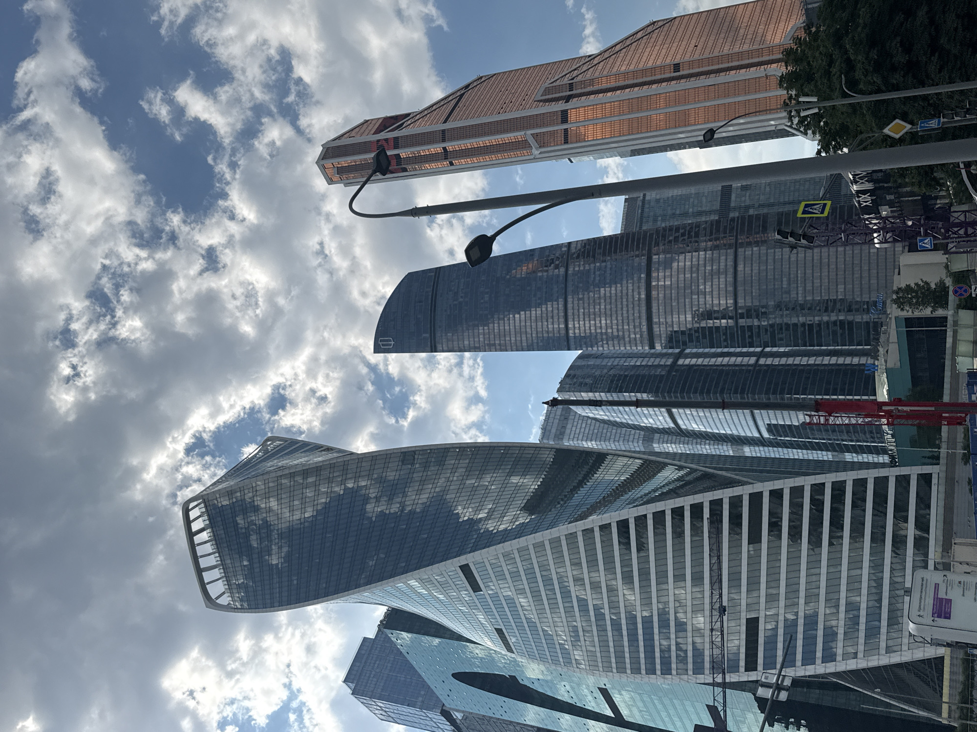 The image captures a low-angle view of several modern skyscrapers in Moscow, Russia, under a cloudy sky. The most prominent building on the left is the twisting "Eurasia Tower" or "SM City Towers," characterized by its unique spiral design and glass facade reflecting the sky and surrounding structures. To its right are other tall buildings with dark, reflective glass surfaces, including the oval-shaped "Naberezhnaya Tower" complex. In the background, the distinctive copper-colored skyscraper "Nakhimov Tower" is visible, its angled facade catching the light. The scene is dominated by the imposing architecture of the Moscow International Business Center (Moscow City). A street lamp with two lights stands in the foreground, along with traffic signals and a pedestrian crossing sign. In the lower part of the image, construction equipment, including a red crane, and trees are visible, suggesting ongoing development or proximity to a green area. The overall impression is one of a contemporary urban landscape with striking architectural designs.