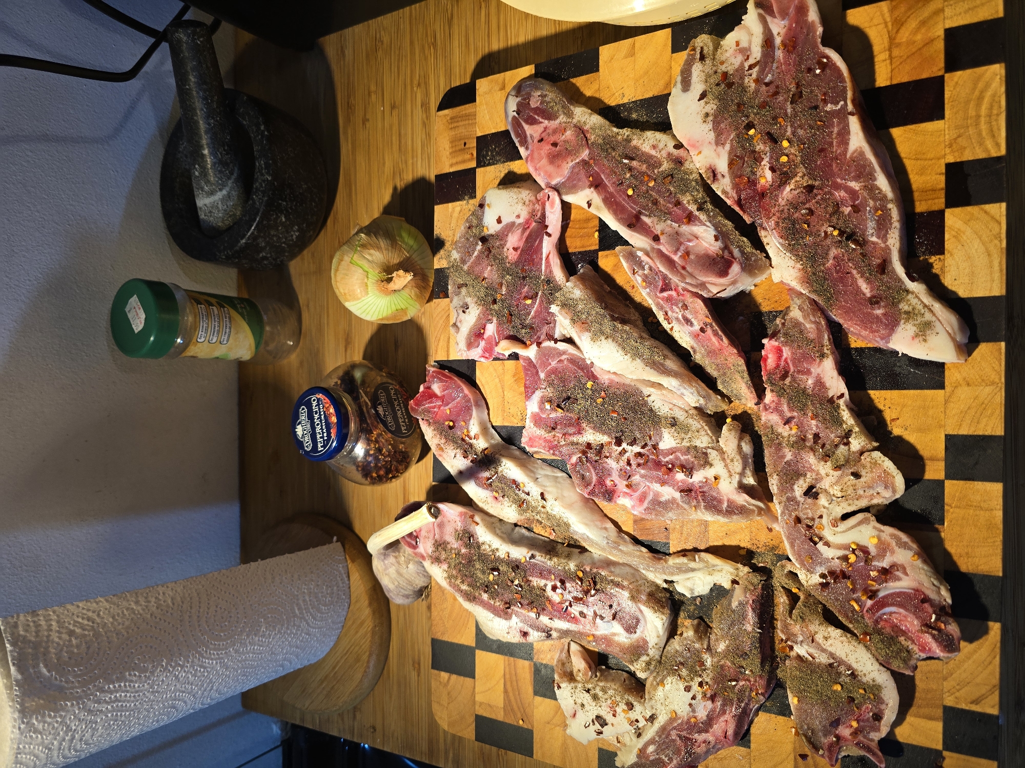 On a checkered wooden cutting board, likely in a kitchen in Even Yehuda, Israel, lie approximately ten seasoned lamb chops. The chops are sprinkled with dried herbs and red pepper flakes. To the left of the cutting board are a grey stone mortar and pestle, a jar of dried spices with a green lid, an onion, and a jar labeled "PEPERONCINO". Below these items is a roll of paper towels. The lighting suggests a kitchen environment. The image captures a culinary preparation scene.